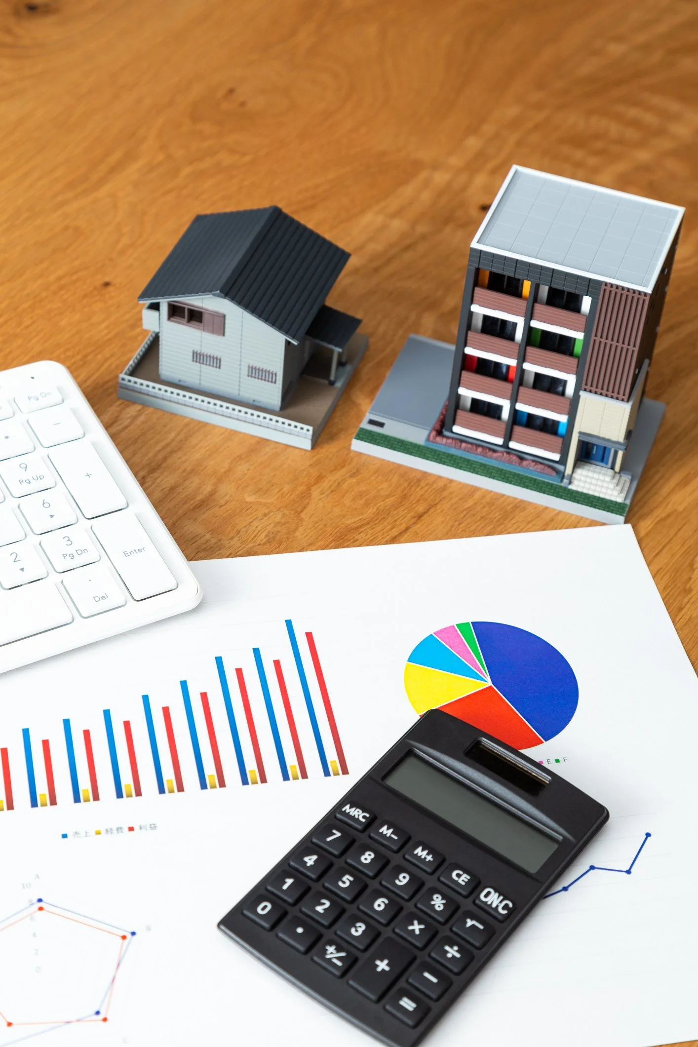 Miniature model of a house and an apartment building placed on a wooden surface with financial documents, a calculator, and part of a computer keyboard.