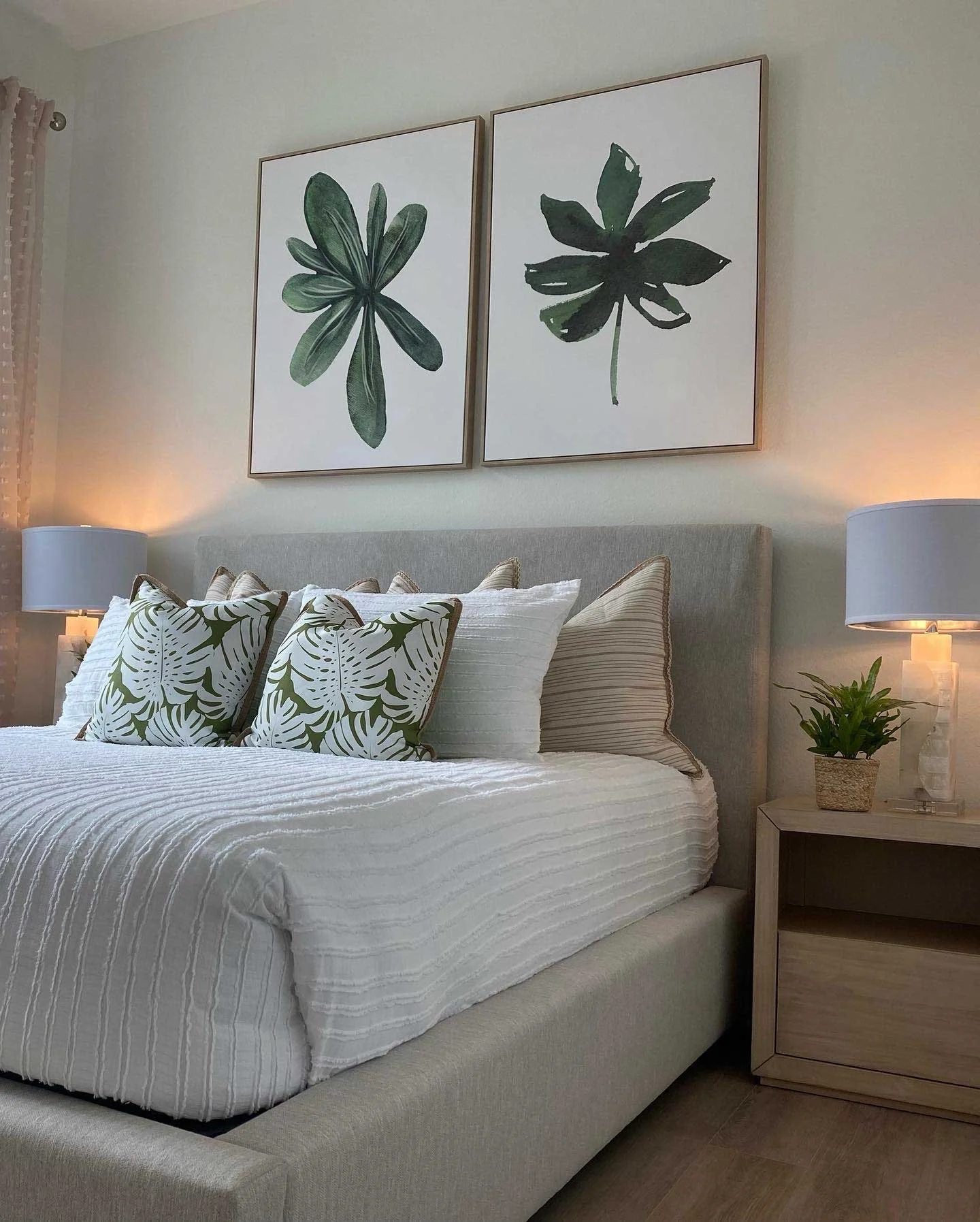Bedroom with a beige upholstered bed, white striped bedding, and decorative pillows with leaf prints. Two nightstands with lamps and a potted plant. Two large framed botanical prints hanging above the bed.
