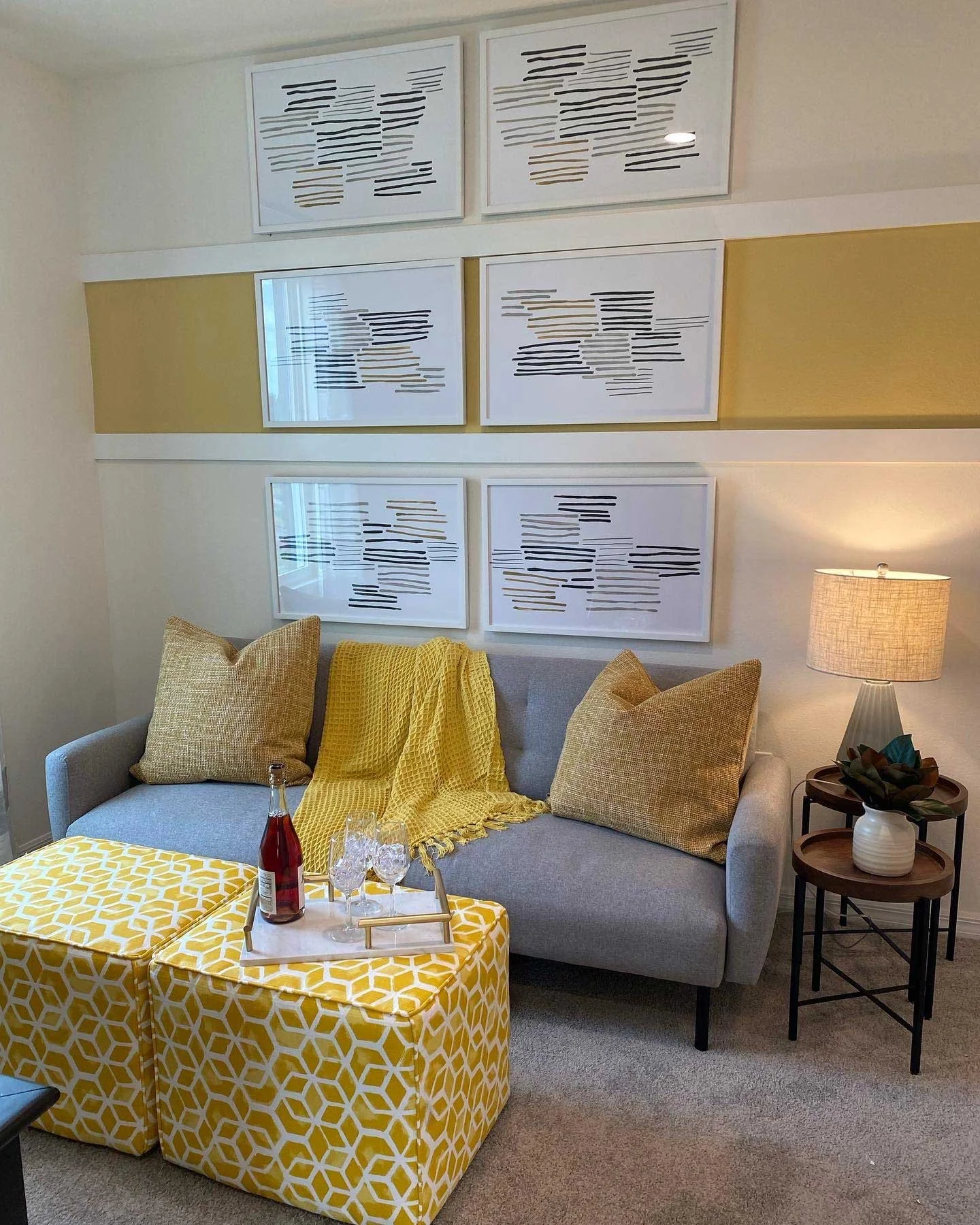 Living room with gray sofa, yellow pillows, yellow throw blanket, yellow patterned ottomans, wall art with abstract black and white lines, side table with lamp and plant, and a tray with drinks.