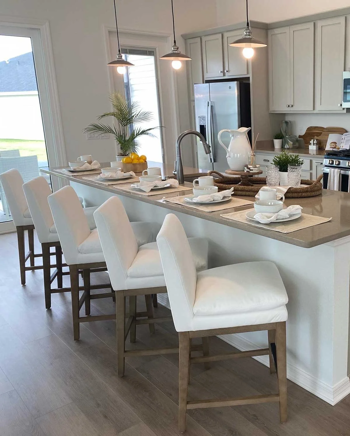 A modern kitchen with a large island, white chairs, and decorative table setting, featuring pendant lights and a view of the outdoors through sliding glass doors.