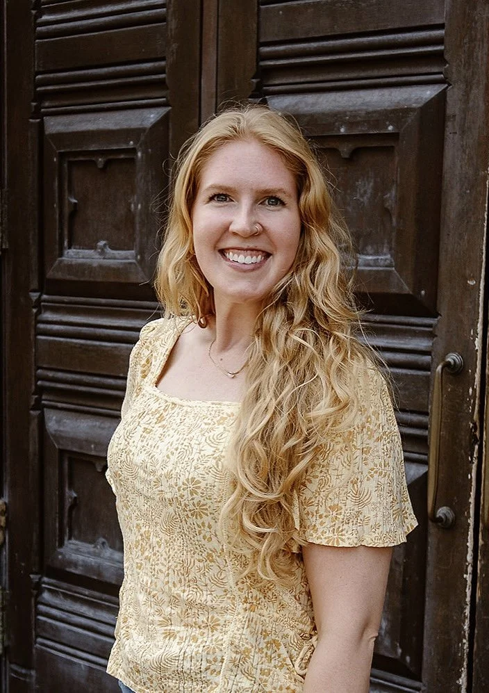 A young woman with long, curly blonde hair and a nose ring smiling in front of a dark wooden door.