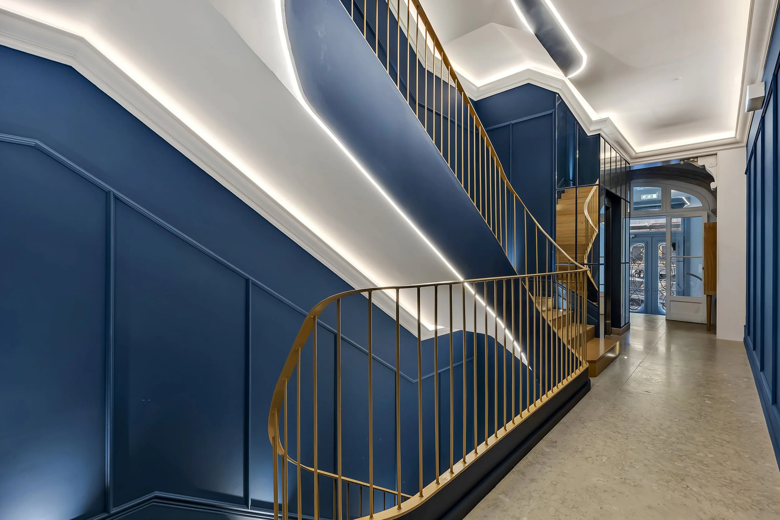 Interior de um prédio com escadas de design moderno, paredes azuis, corrimãos dourados e iluminação embutida.