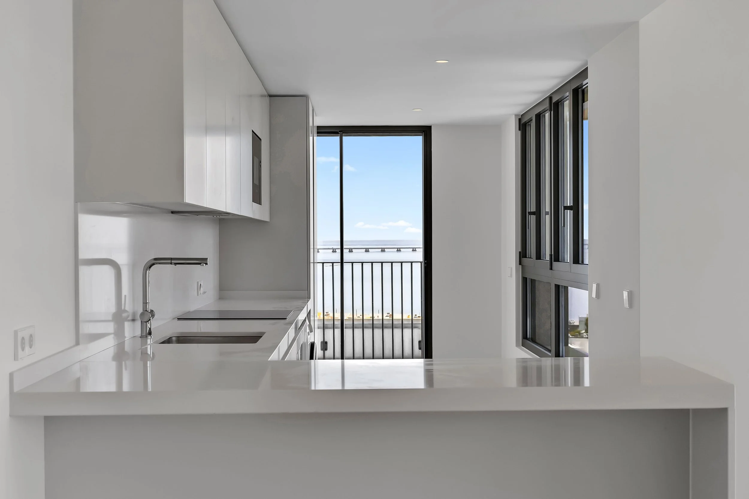 Cozinha moderna com balcão de mármore branco, pia, armários brancos, vidro para varanda com vista para o céu e o mar, janelas grandes com moldura escura.