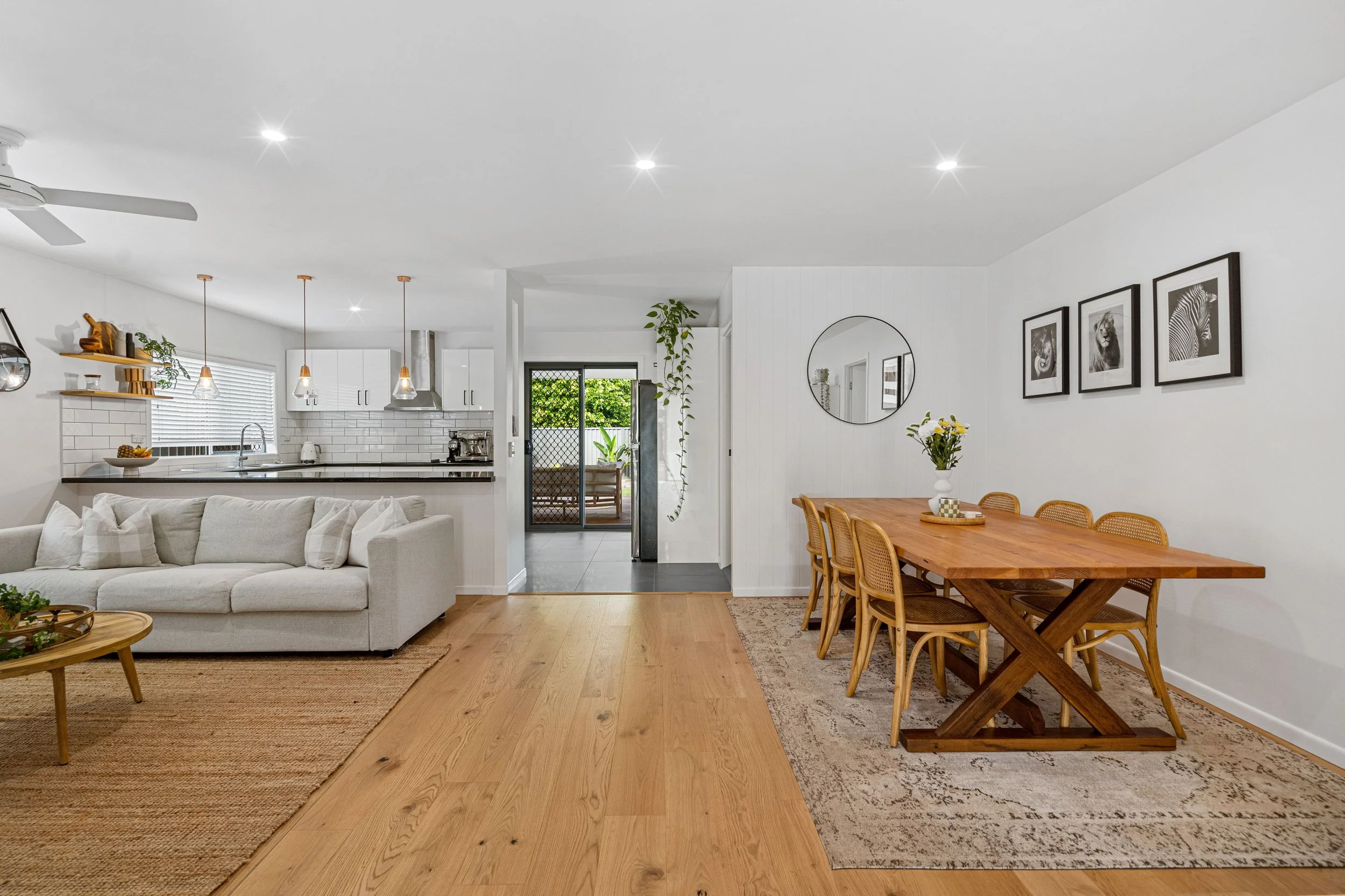 Sala de jantar com mesa de madeira e oito cadeiras, parede branca com quadros preto e branco, sala de estar com sofá branco, cozinha com armários brancos e balcão de granito.