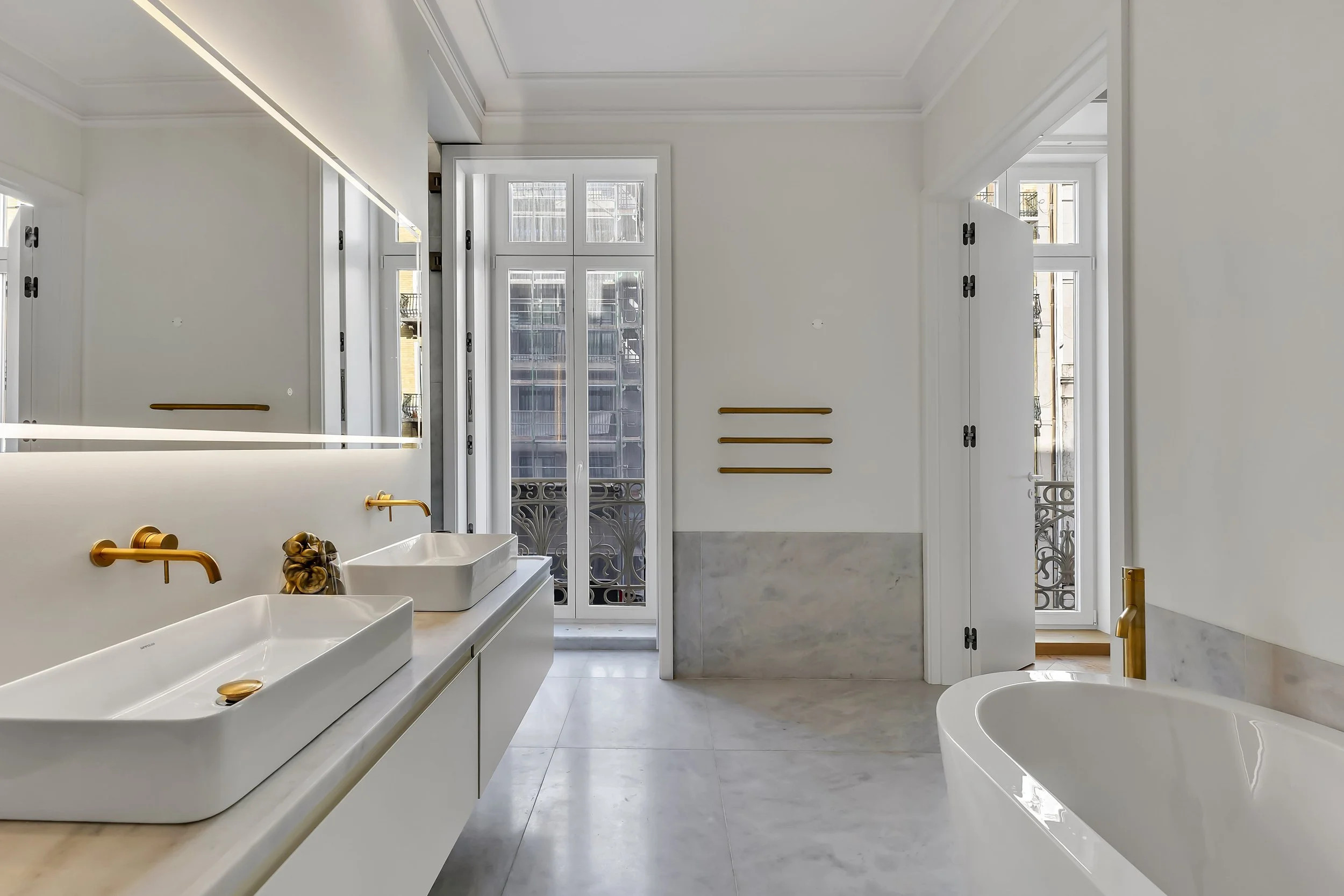 Banheiro com duas pias retangulares brancas com torneiras douradas, espelho grande, janela com vista para edifícios e varanda, banheira branca, paredes brancas, detalhes em mármore e acessórios dourados.