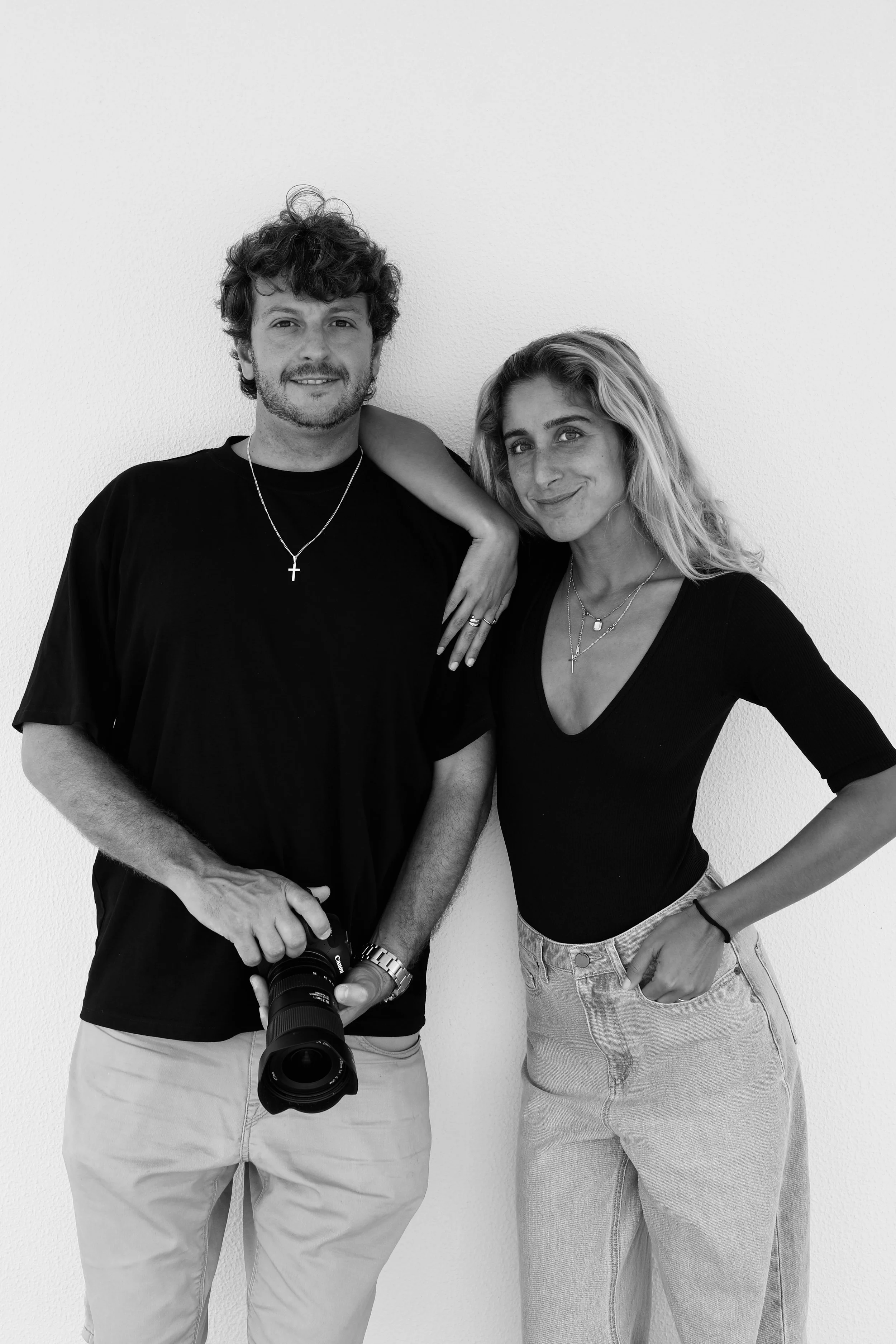 Homem e mulher posando juntos contra parede branca, ele segura uma câmera fotográfica, ambos usam roupas pretas e acessórios com cruz.
