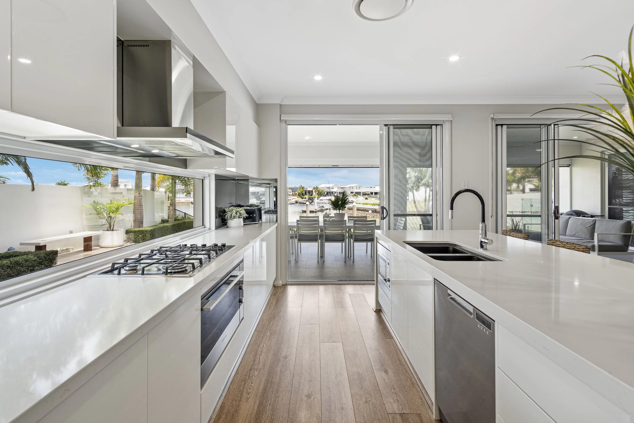 Cozinha moderna com bancada branca, pia preta, vista para uma área externa com mesa e cadeiras, e janelas com vista para uma piscina e jardim