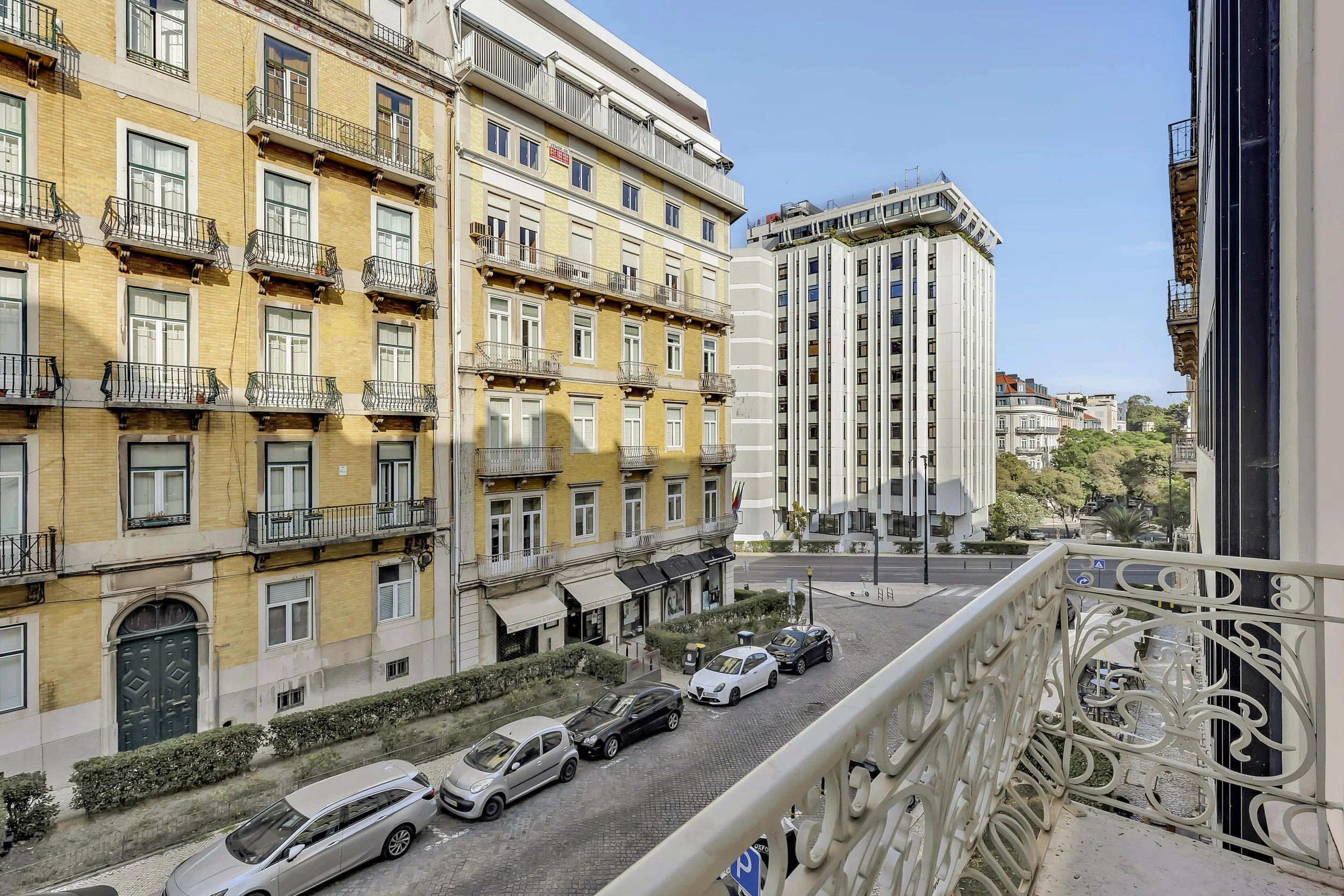 Vista de rua urbana com edifícios residenciais e comerciais, carros estacionados na calçada, árvores, e céu claro, tirada de um varanda com grade ornamental.