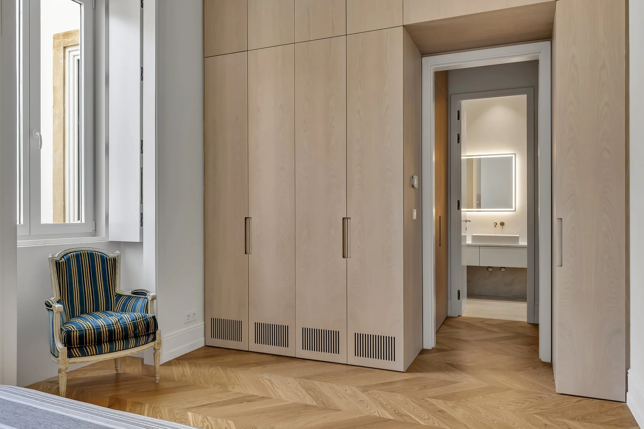 Quarto com piso de madeira, armários de roupas de cor clara, uma cadeira decorativa com tecido listrado azul e amarelo, janela aberta com vista para uma parede de pedra, e um banheiro ao fundo com espelho retangular com luz.
