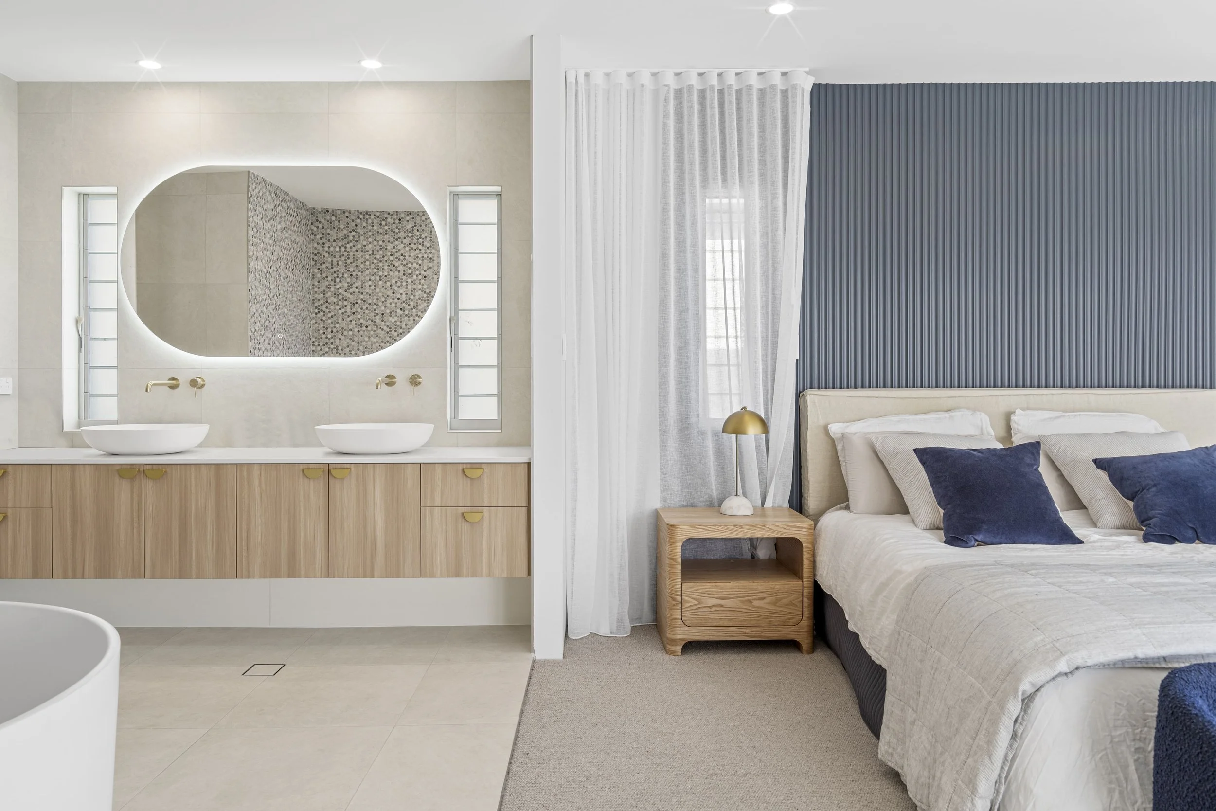 Quarto de casal com cama de roupas brancas, almofadas azul escuro, cabeceira branca, parede de gesso azul com detalhes ripados, cortina branca translúcida, ao lado uma mesa de cabeceira de madeira com luminária dourada, e ao fundo um banheiro com esp
