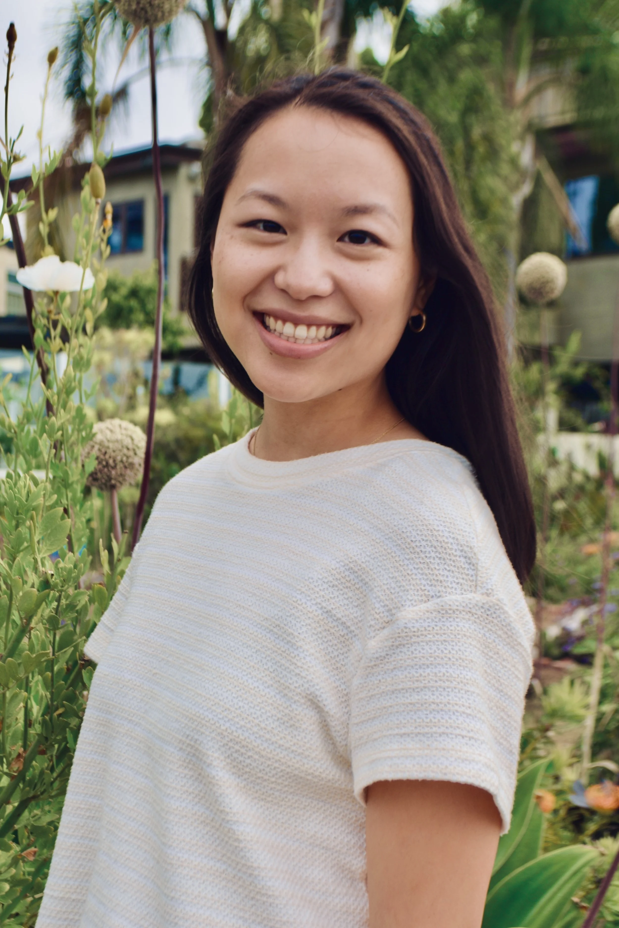 A smiling woman with dark hair and small hoop earrings standing outdoors among green plants and flowers, with a house in the background.