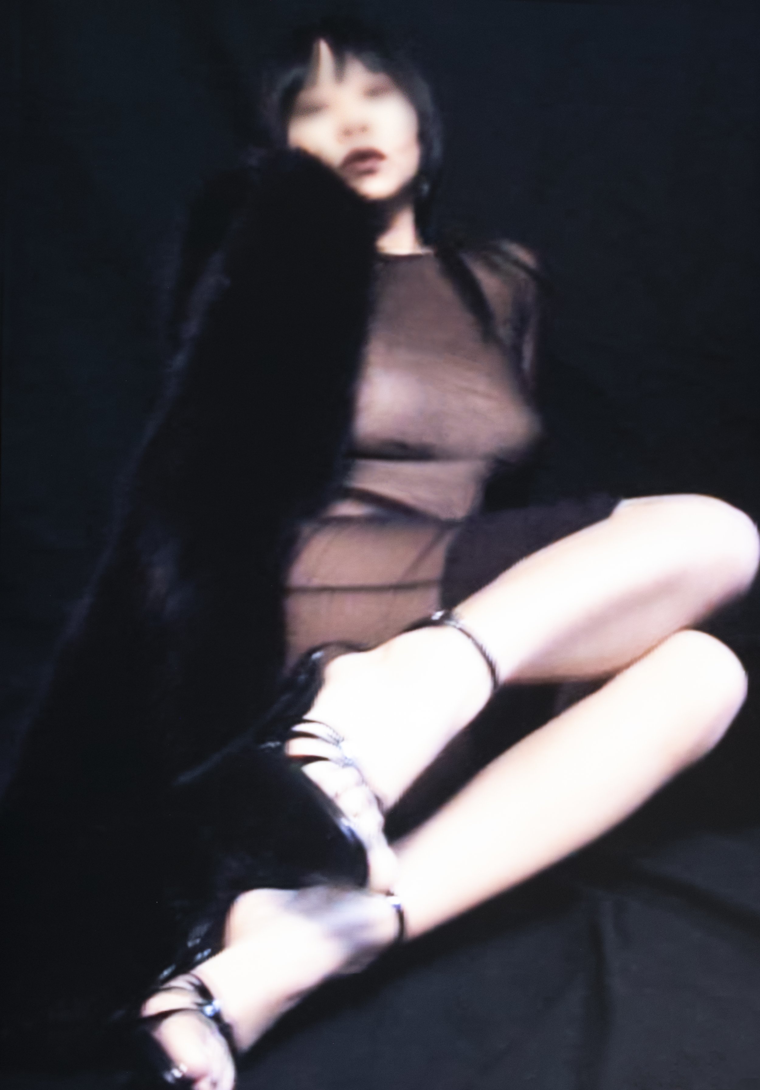 NYC Dominatrix Saturn Veil wearing a sheer black top and black high heels, sitting with her legs crossed against a dark background.