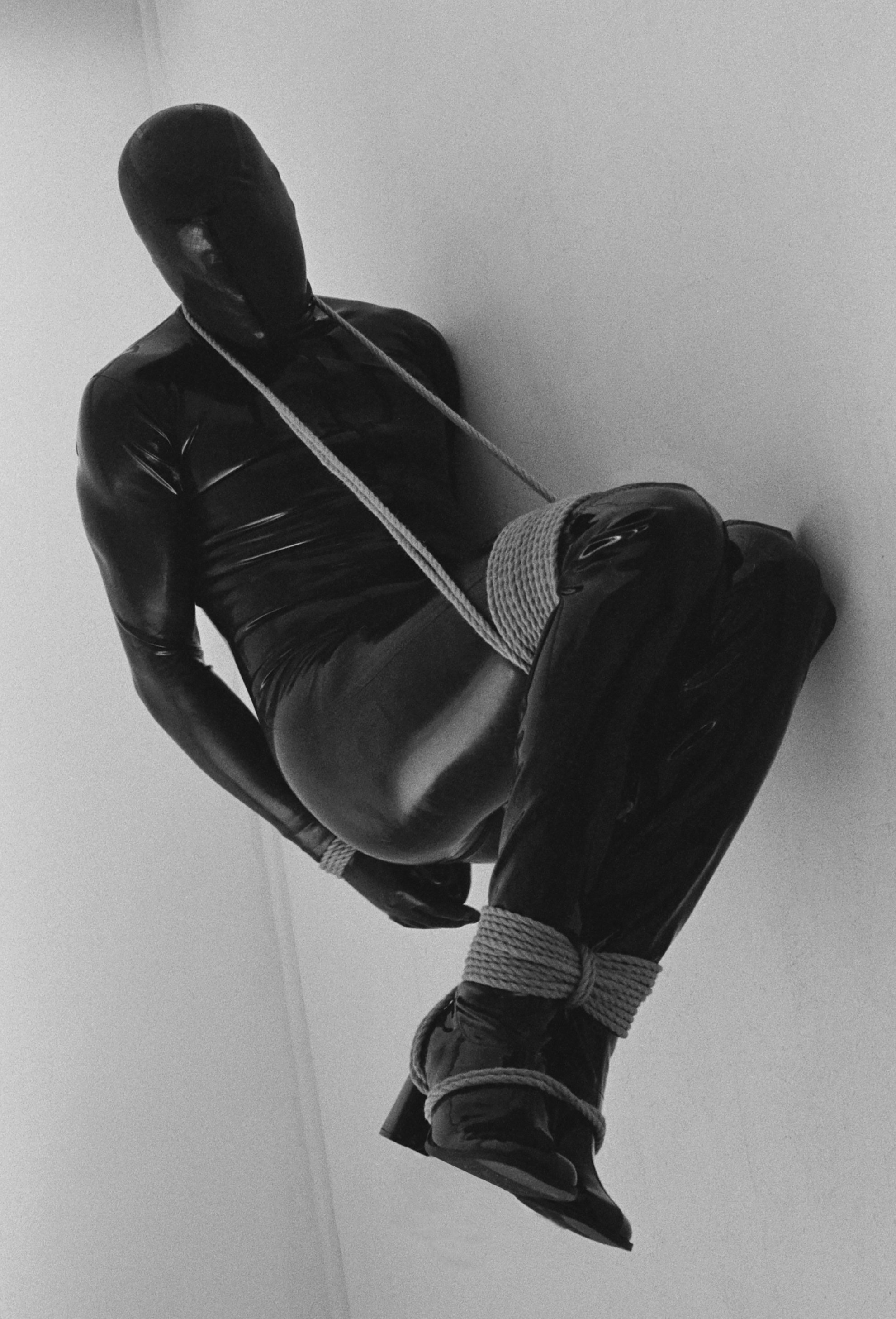 Person in black latex outfit tied with rope sitting against a wall in a minimalist room.