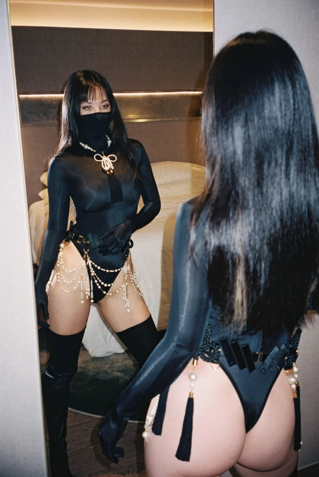 Lady Eden dressed in black lingerie with pearl chains and accessories, wearing a mask, looking at her reflection in a mirror.