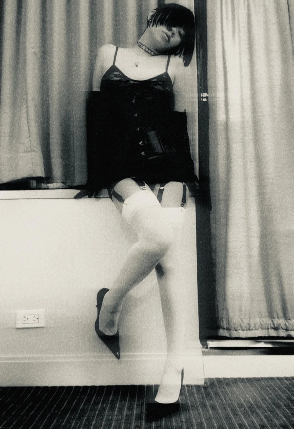 SF Dominatrix Saturn Veil wearing a choker, a black dress with lace details, thigh-high stockings, and high heels, leaning against a wall with curtains in the background.