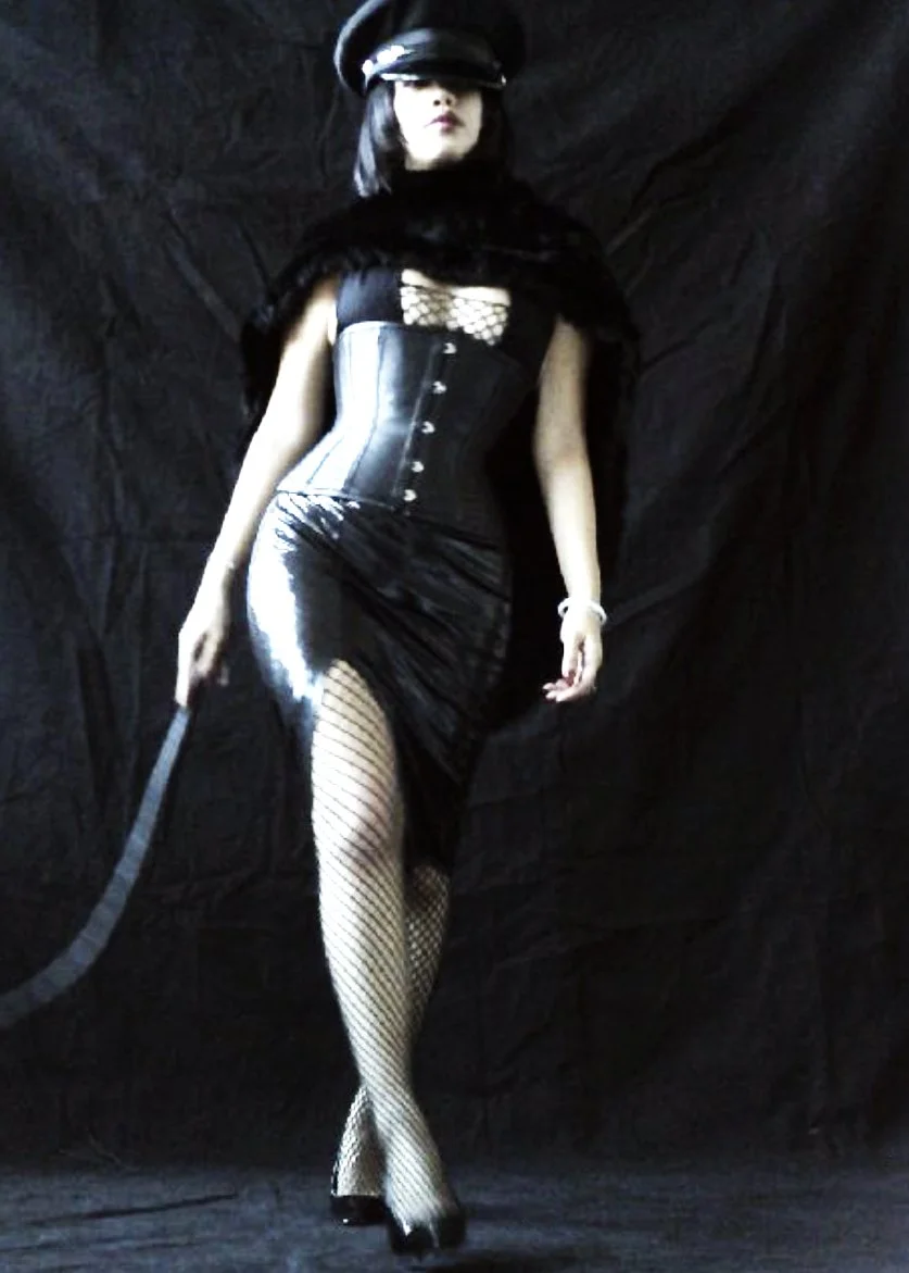 NYC Dominatrix Saturn Veil in black latex and leather with fishnet stockings, wearing a police-style hat, standing against a black background.