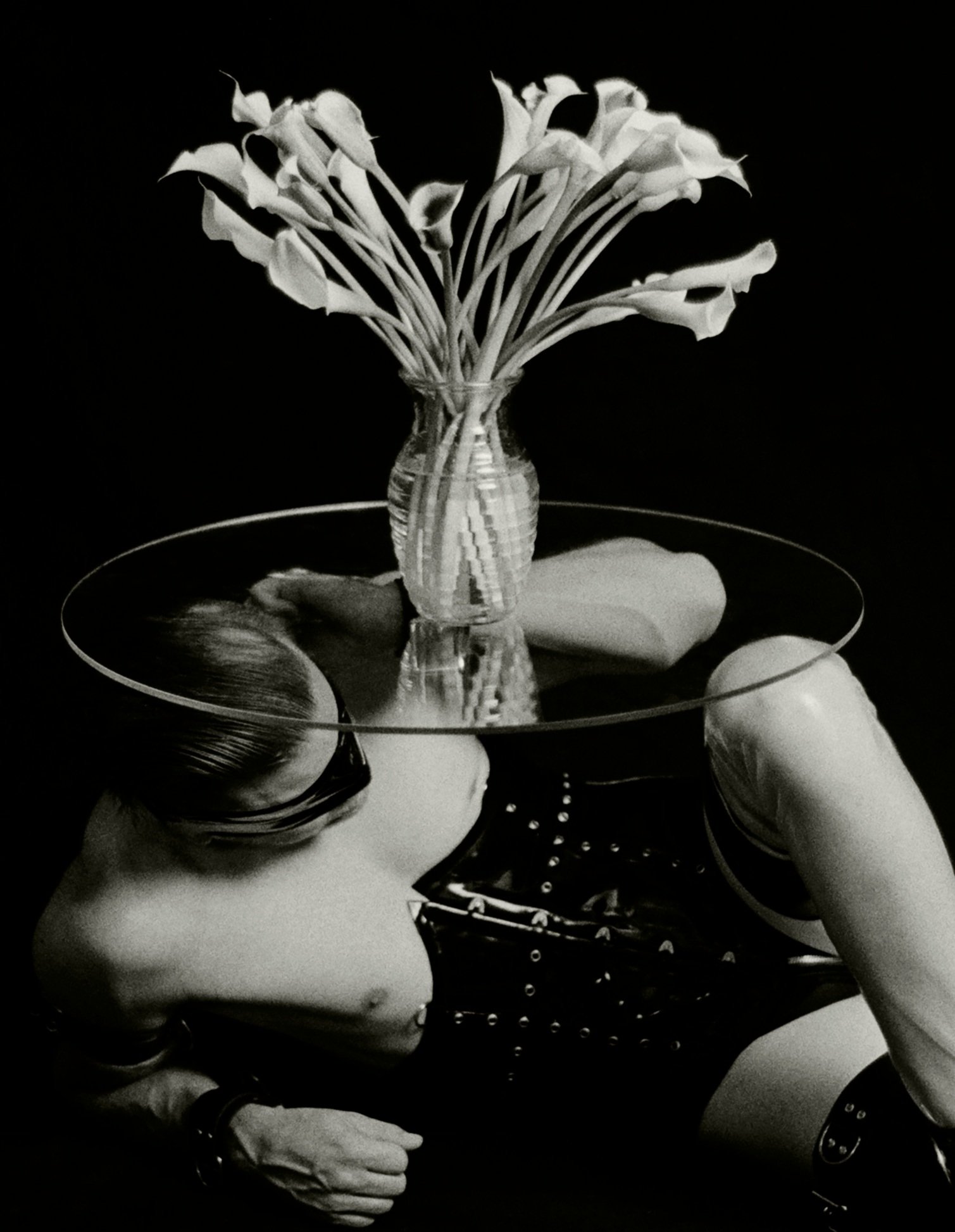 Black and white artistic photo of a person wearing leather clothing, lying beneath a circular glass tabletop with a vase of flowers on top.