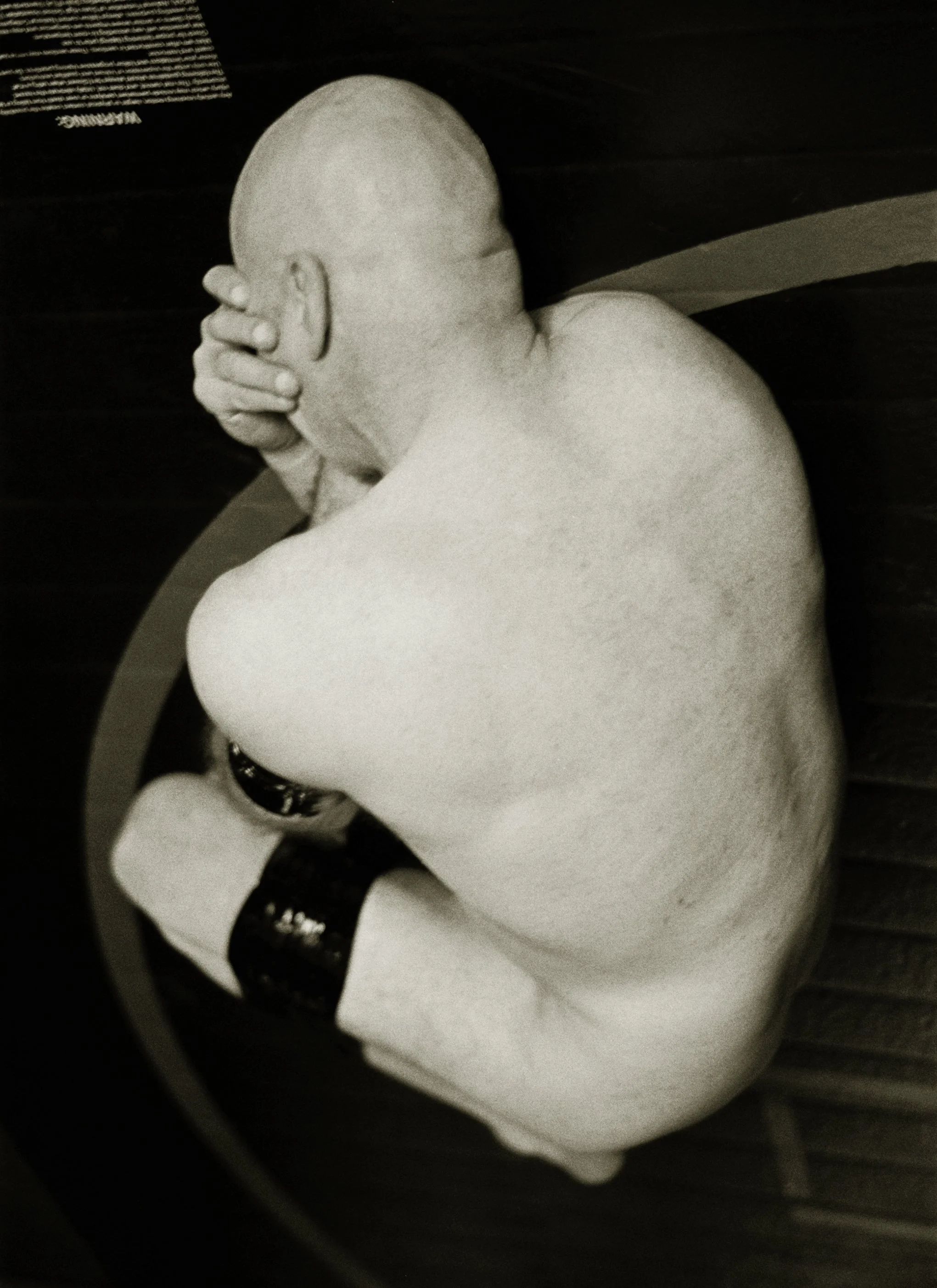 NYC and SF Dominatrix Saturn's Black and white photo of a tattooed, shirtless, bald man sitting on a curved surface with his head resting in his hand and his eyes closed.