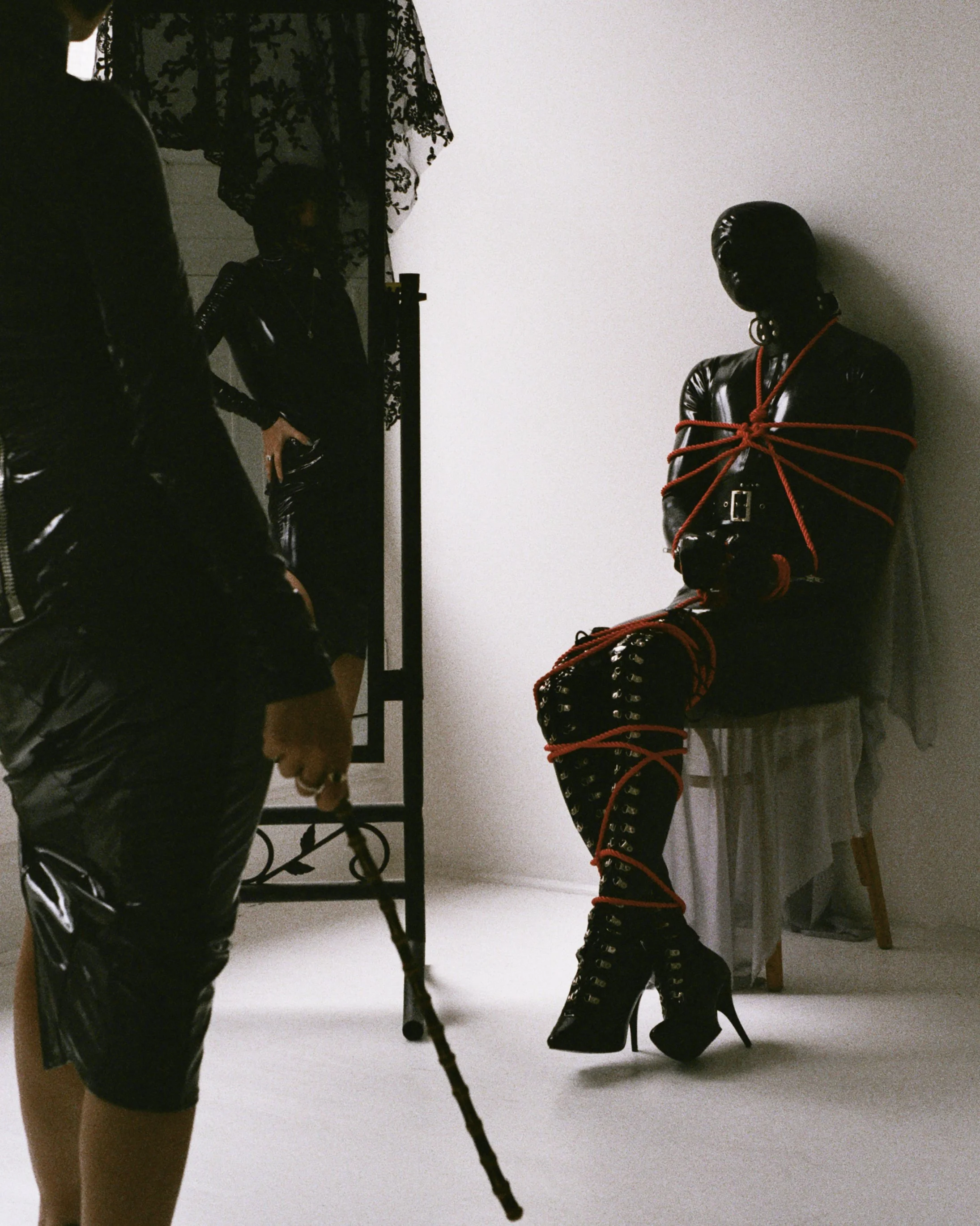 The dim light of the room clings to My black PVC outfit, an almost second skin. I feel the cool, familiar weight of My cane in My hand, and extension of My will. Before Me, he waits—My submissive for the day, bound in coils of vivid red rope atop of 