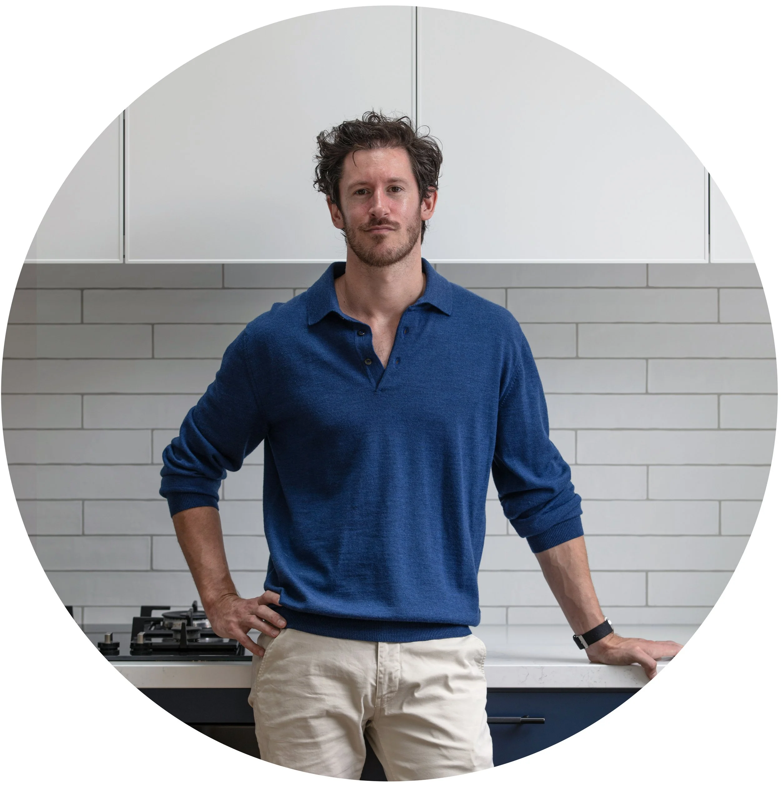 A man standing in a modern kitchen, wearing a blue sweater and beige pants, with one hand on his hip and the other resting on a counter, in front of white subway tile backsplash and a stove.