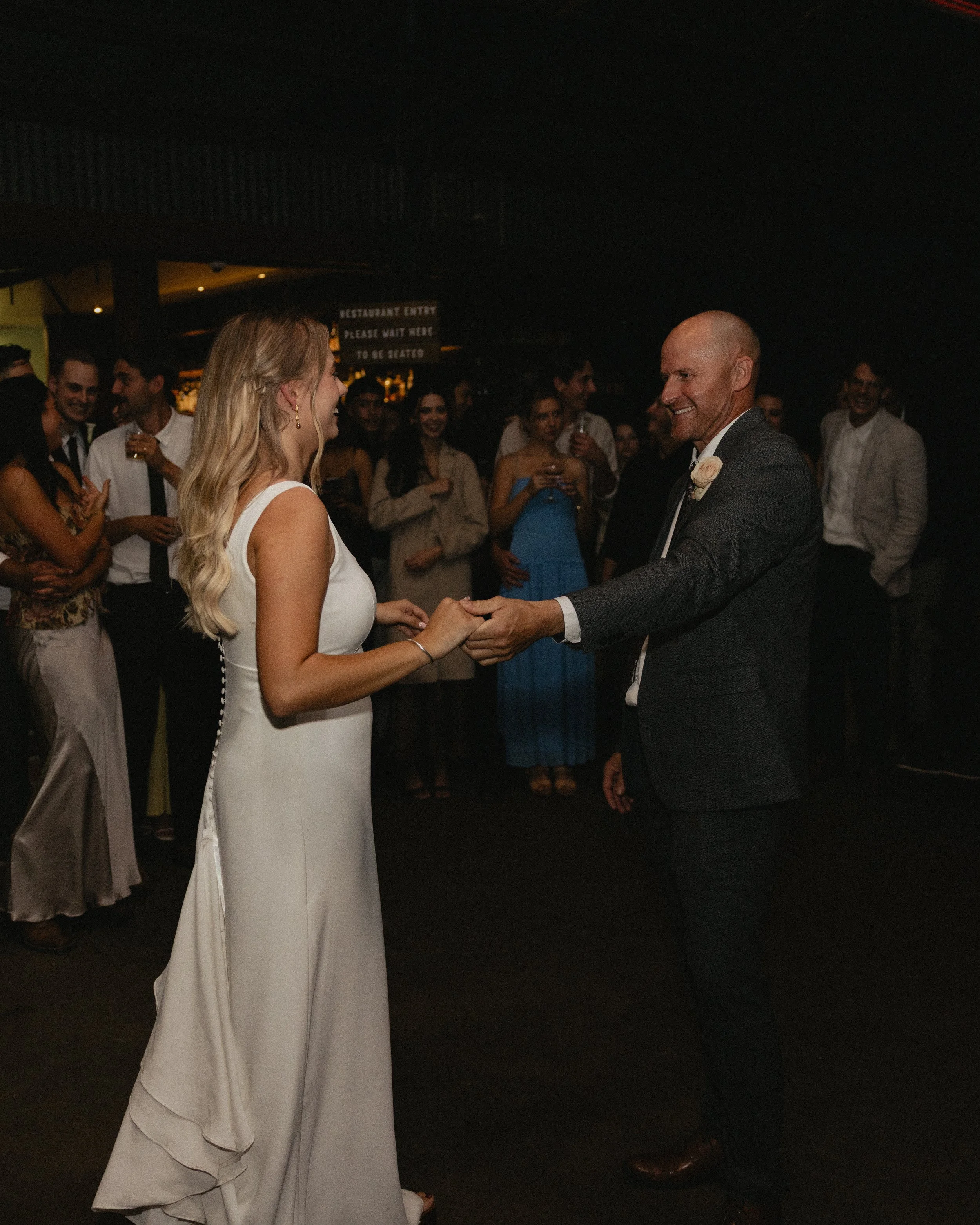 A bride and groom are dancing together at their wedding reception, surrounded by guests in a dimly lit venue.