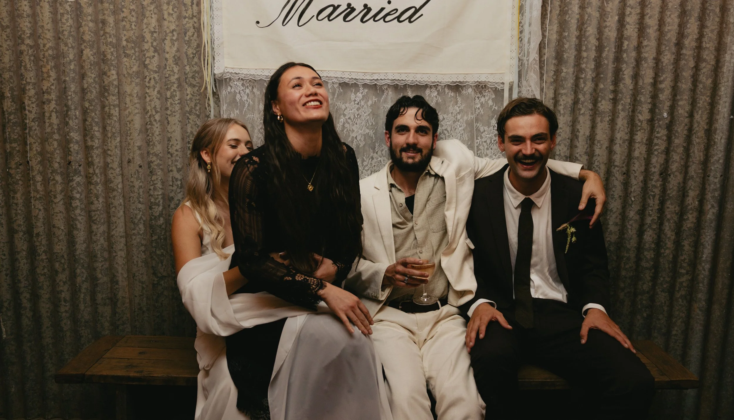 Group of five friends sitting on a wooden bench at a wedding celebration, with a 'Married' banner behind them, smiling and enjoying drinks.