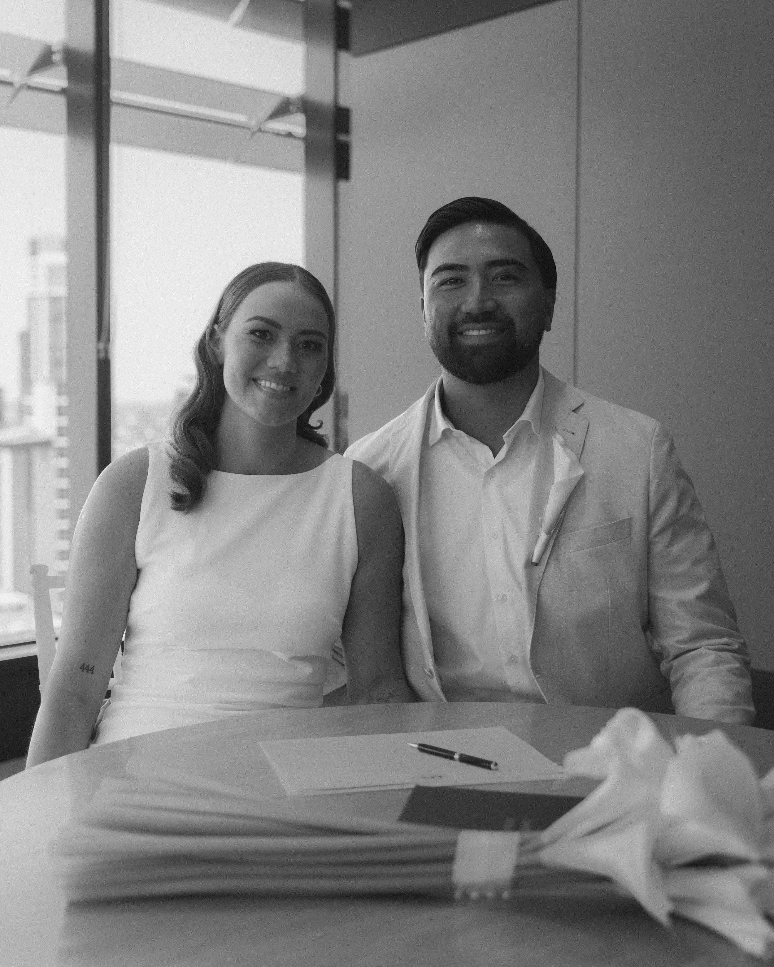 A man and woman in formal attire sitting at a table, smiling at the camera, with documents and a pen in front of them, in an office setting.