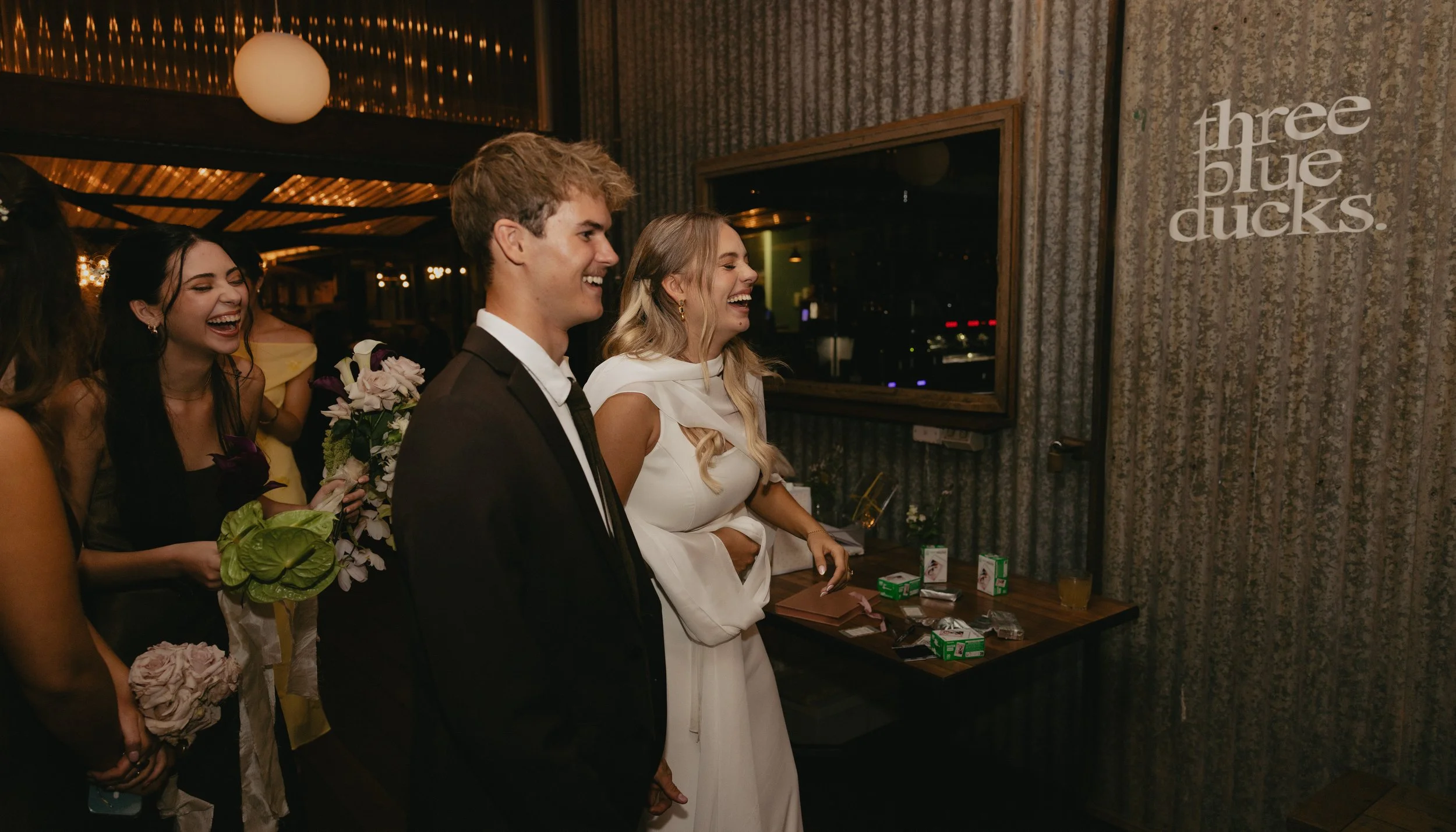 A group of people at a celebration, with some holding bouquets of flowers, laughing and smiling inside a decorated venue with dim lighting and a sign that says 'three blue ducks' on the wall.