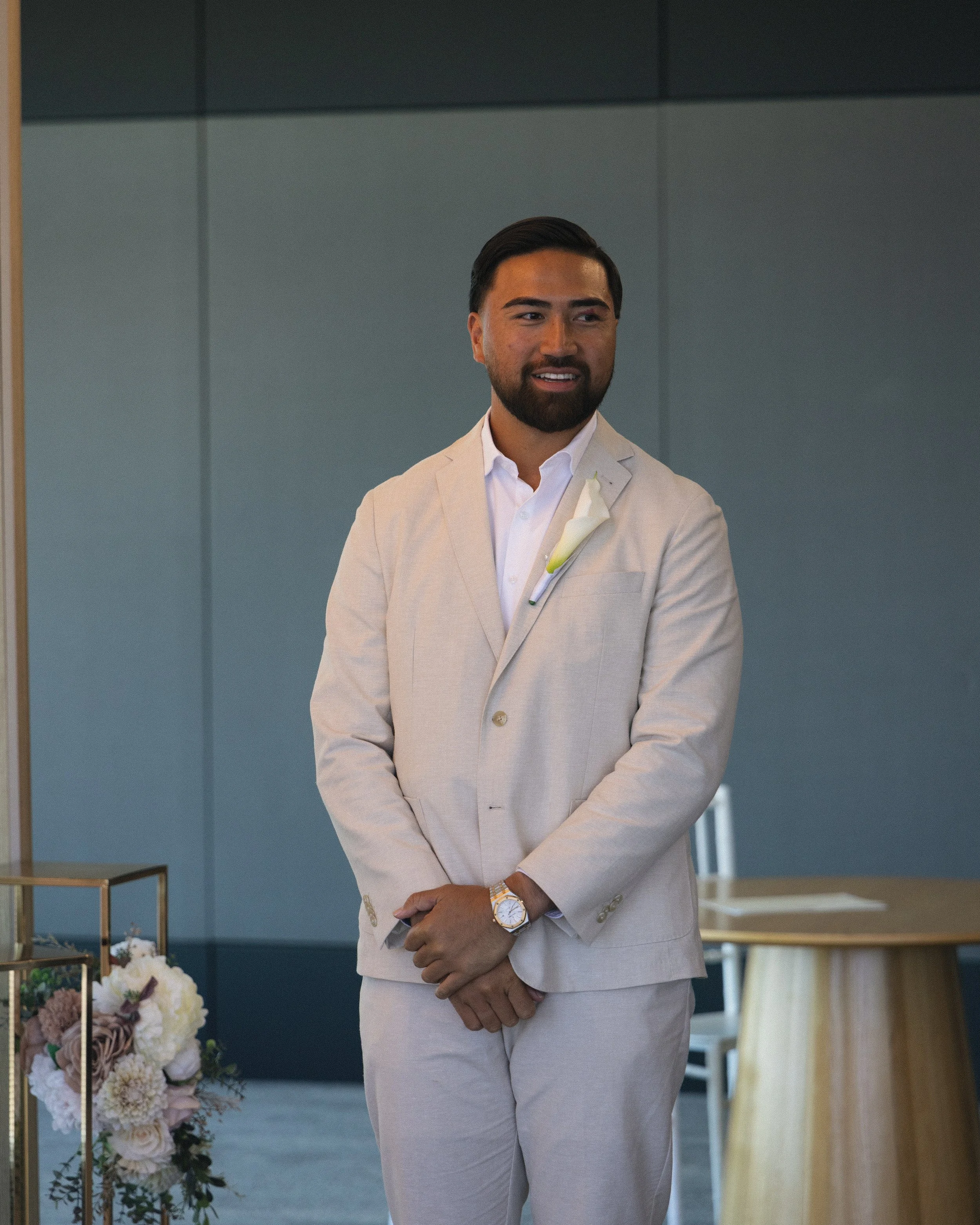 A man in a light-colored suit with a white shirt and a floral lapel pin stands with his hands clasped in front of him, smiling during a formal event in a modern indoor setting.