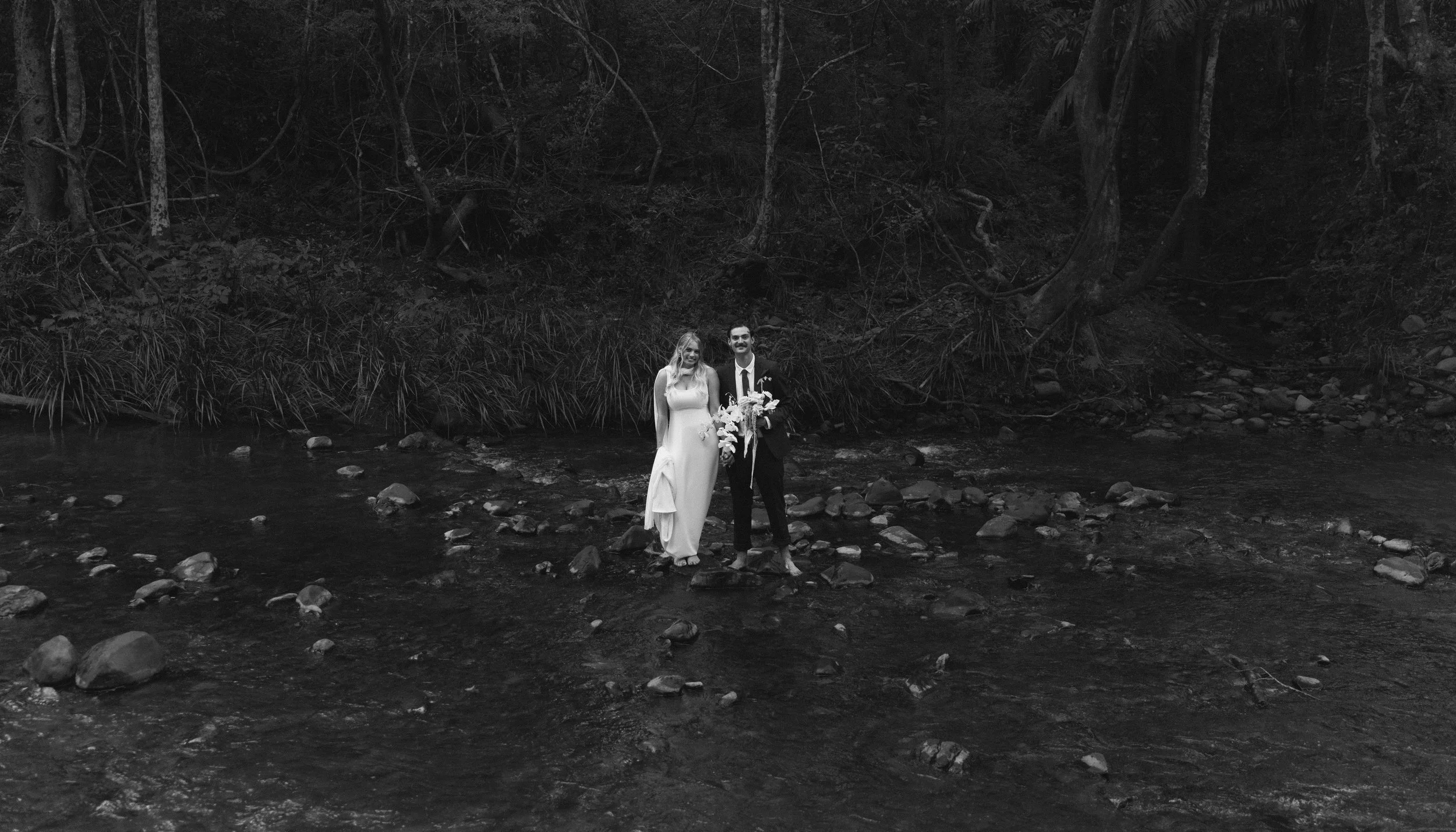 A black and white photo of a bride and groom standing on rocks in a shallow stream surrounded by trees and dense forest.