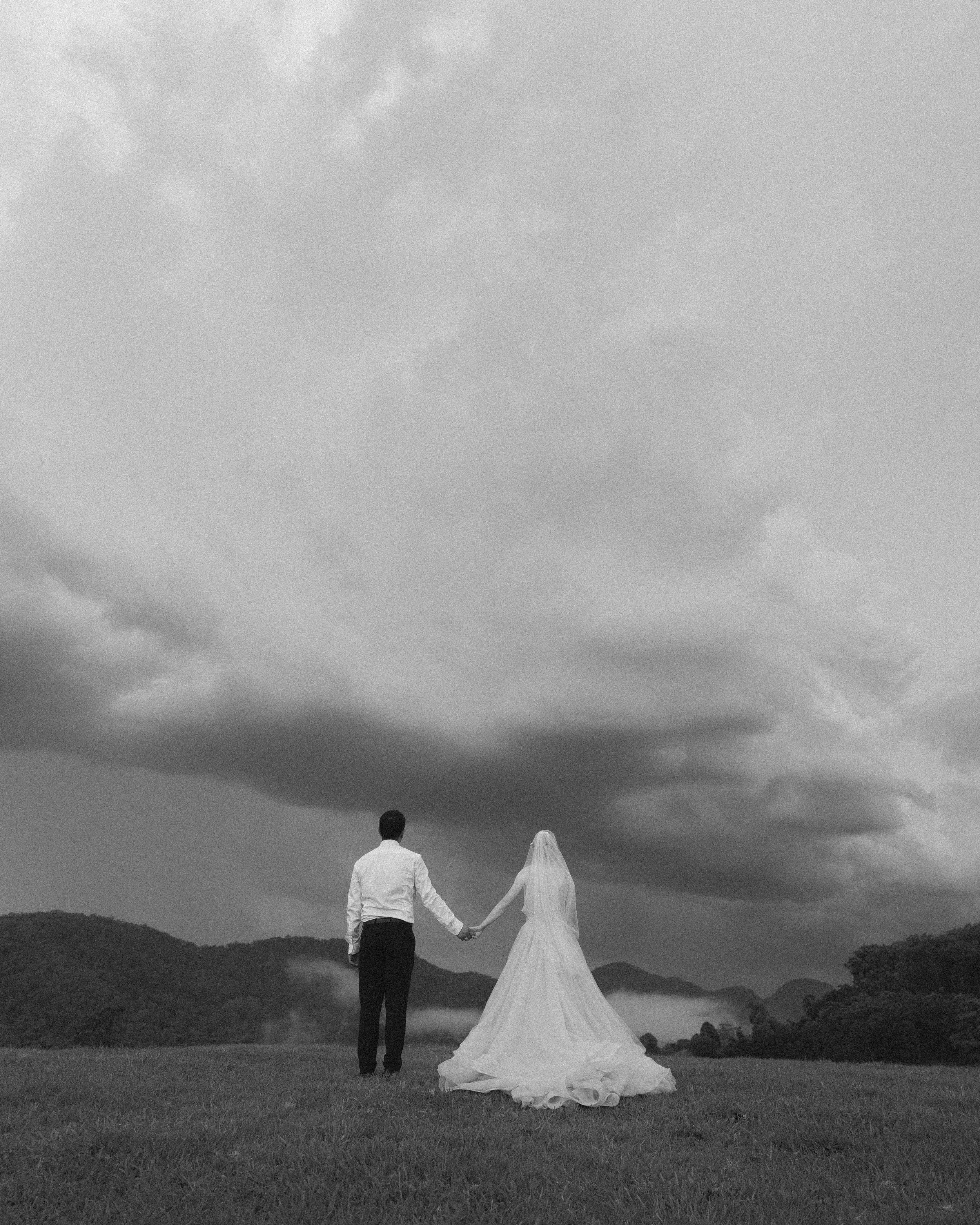 A bride and groom holding hands in a field during a cloudy day with mountains in the background, black and white photograph.