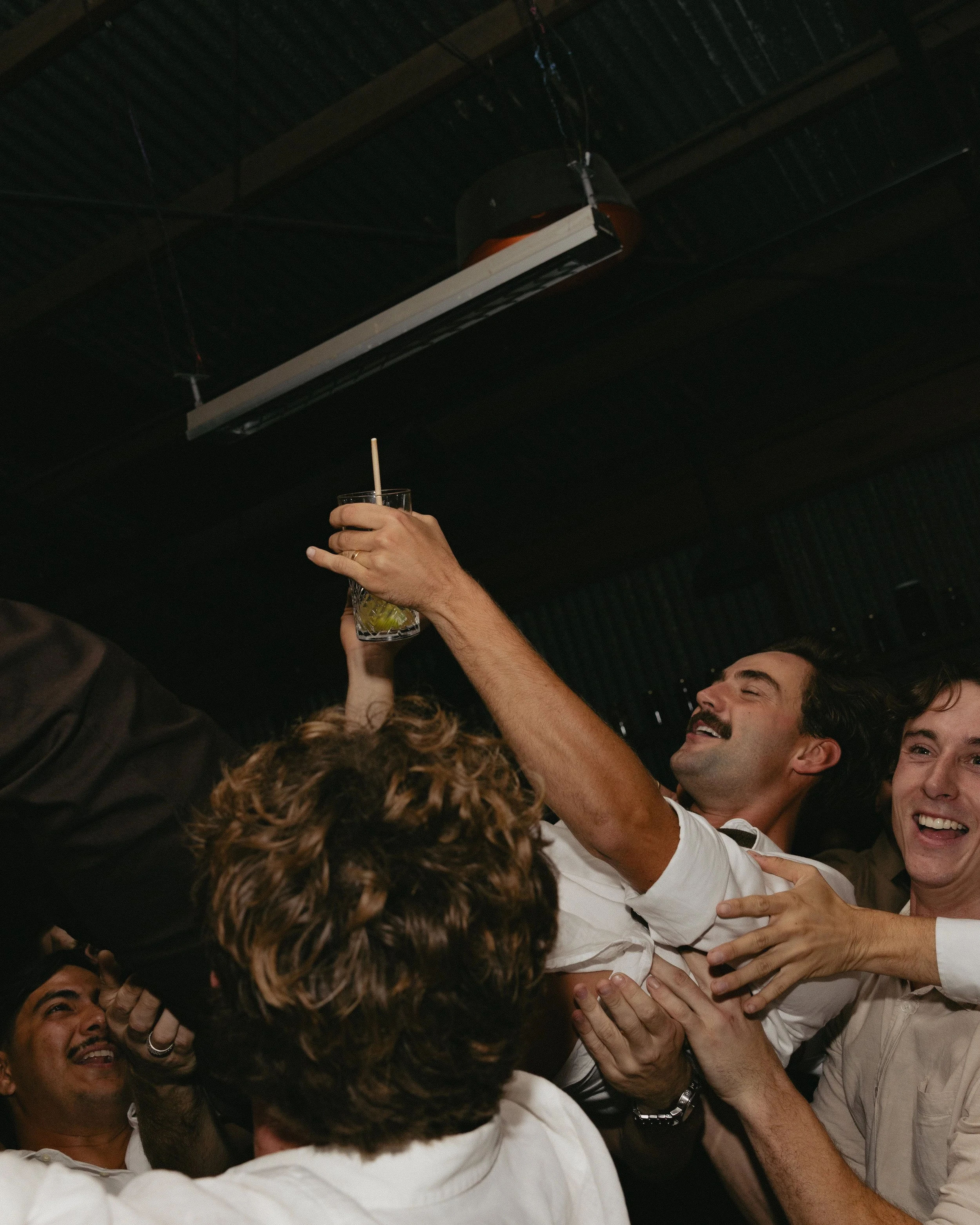 Group of men celebrating, one raising a glass with a straw in it, in a dark indoor setting.