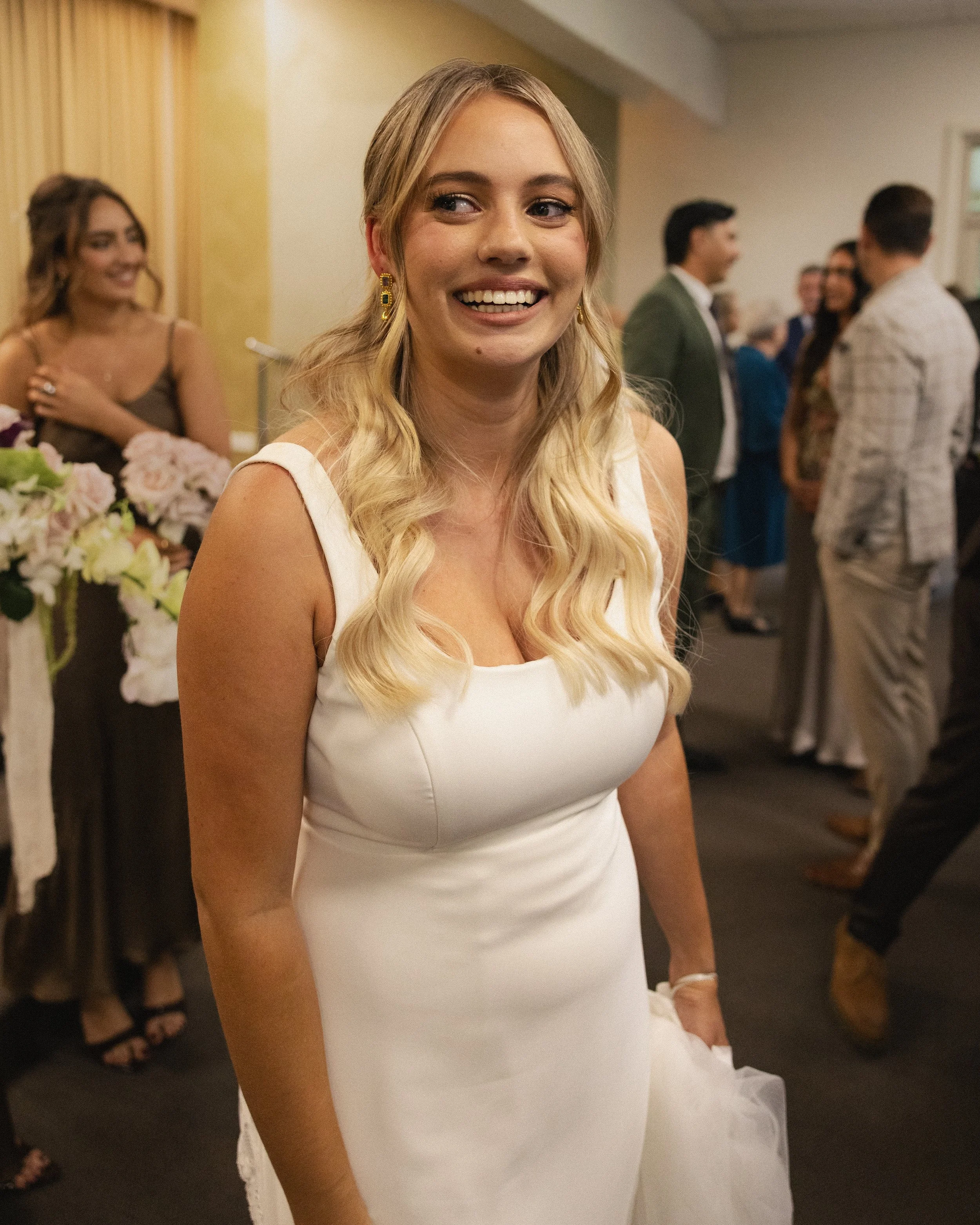 A young woman in a white dress smiling at a social gathering or celebration, with other people in the background.