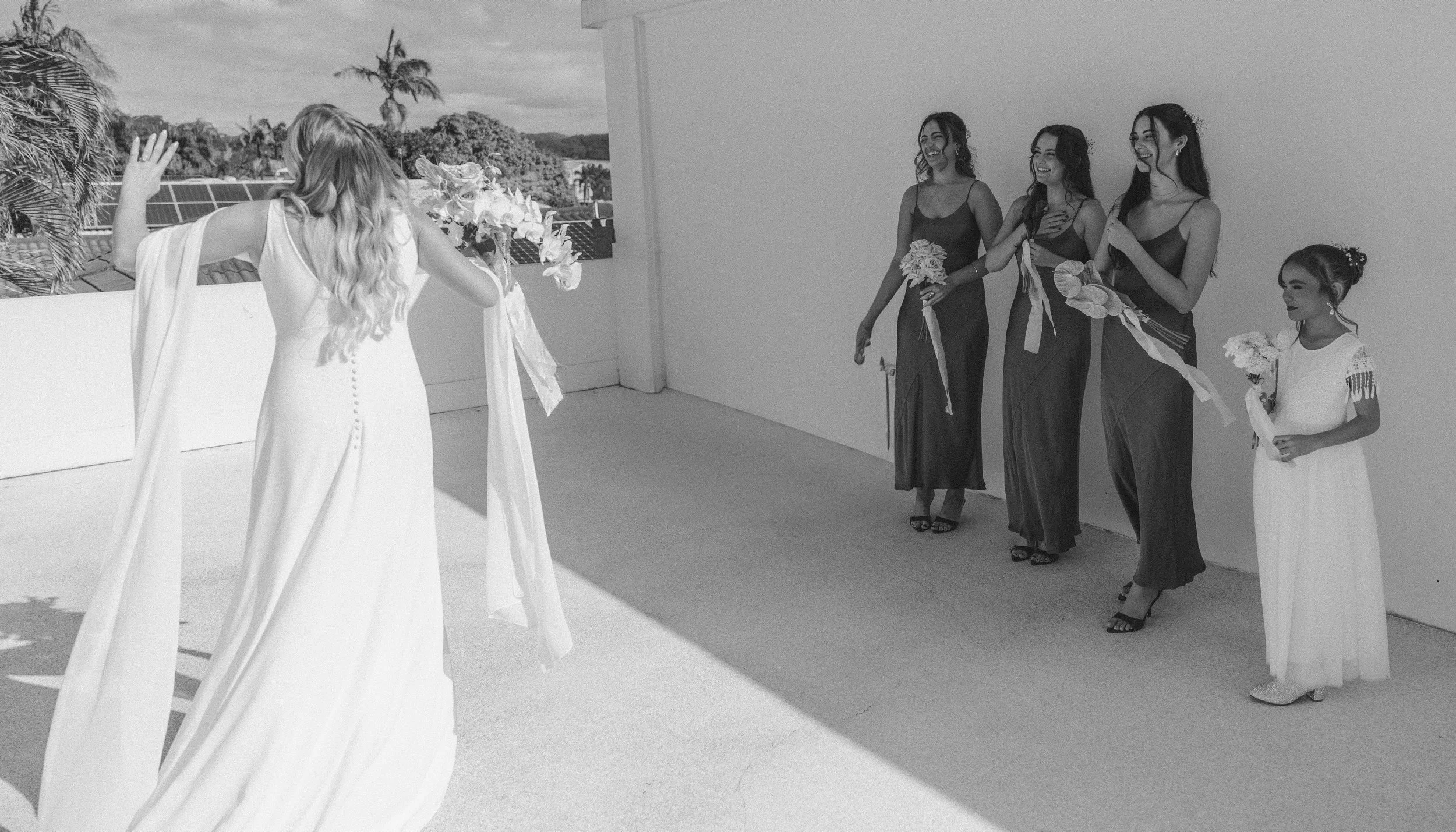 A bride in a white gown with buttons down the back faces four women and a girl, who are holding bouquets and smiling, on a rooftop terrace with a cityscape and trees in the background.