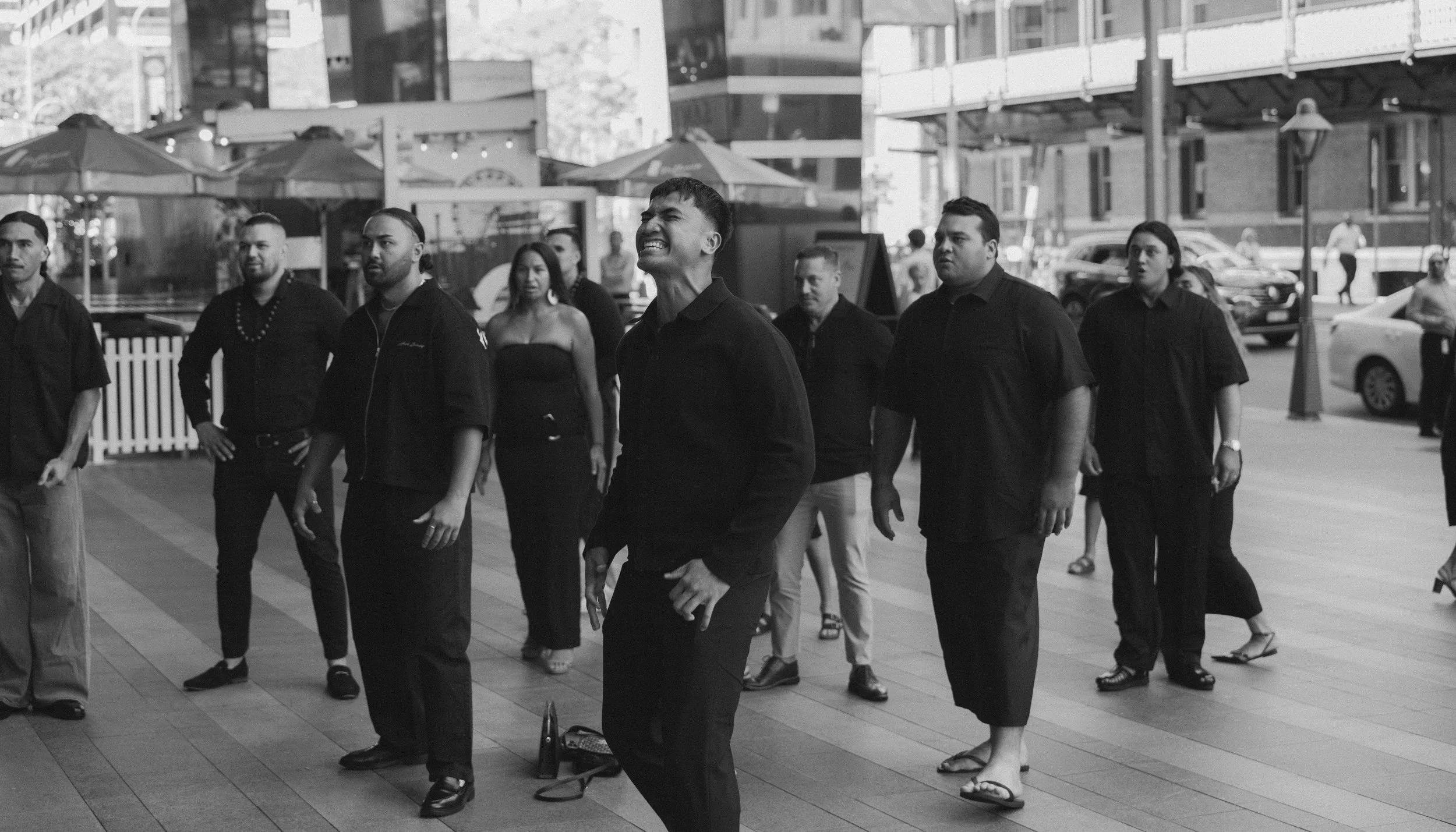 A group of people dressed in black, standing on a city sidewalk, with some wearing traditional Asian attire. One man in the center is smiling and appears to be dancing or performing, with others looking on.