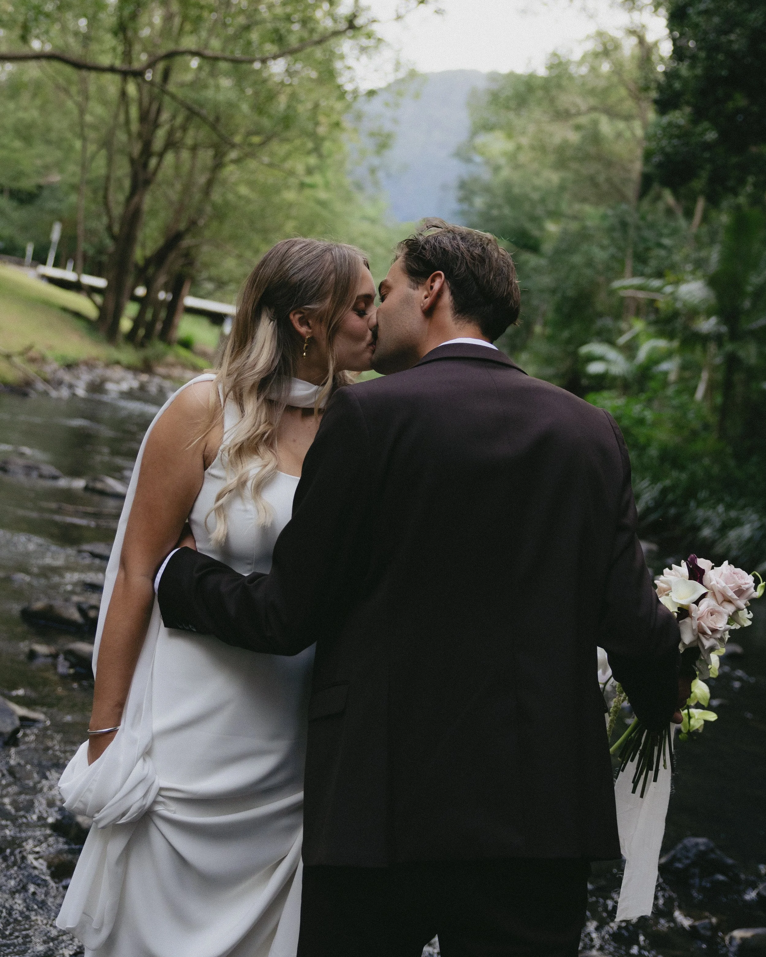 A couple in wedding attire kissing by a stream in a lush green forest, with mountains in the background.