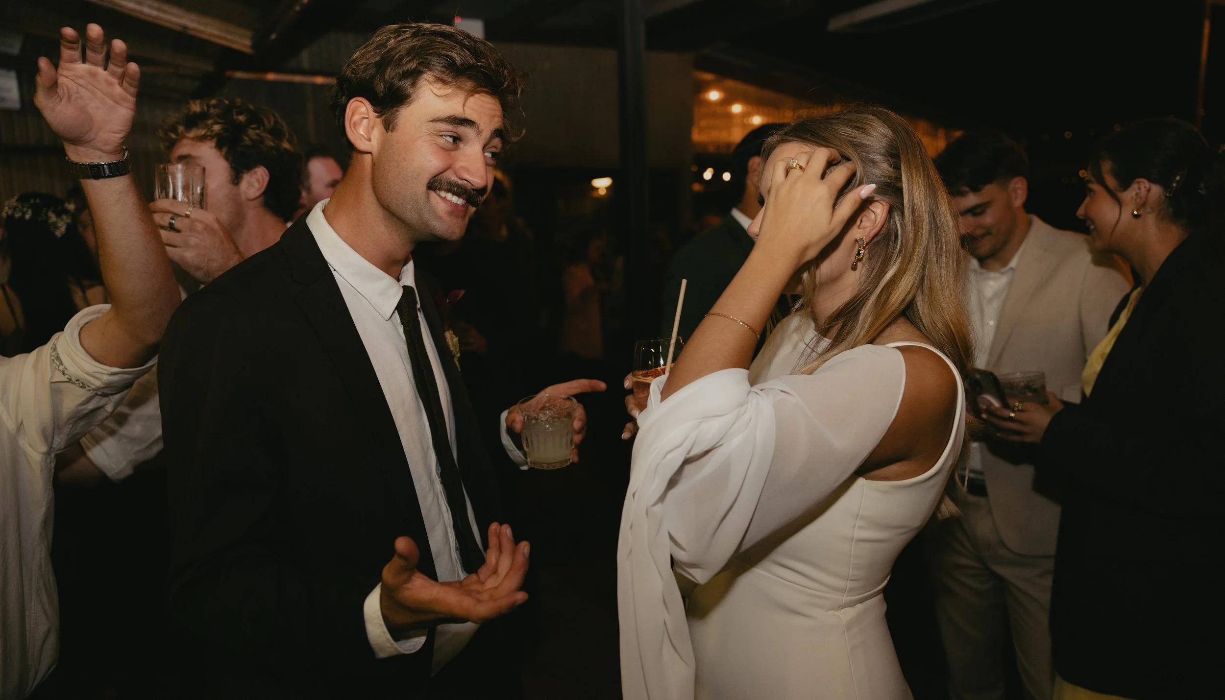 A man and woman are talking and laughing at a social gathering or party, with drinks in their hands. The man has dark hair and a mustache, dressed in a black suit, and the woman has blonde hair and is wearing a white dress.