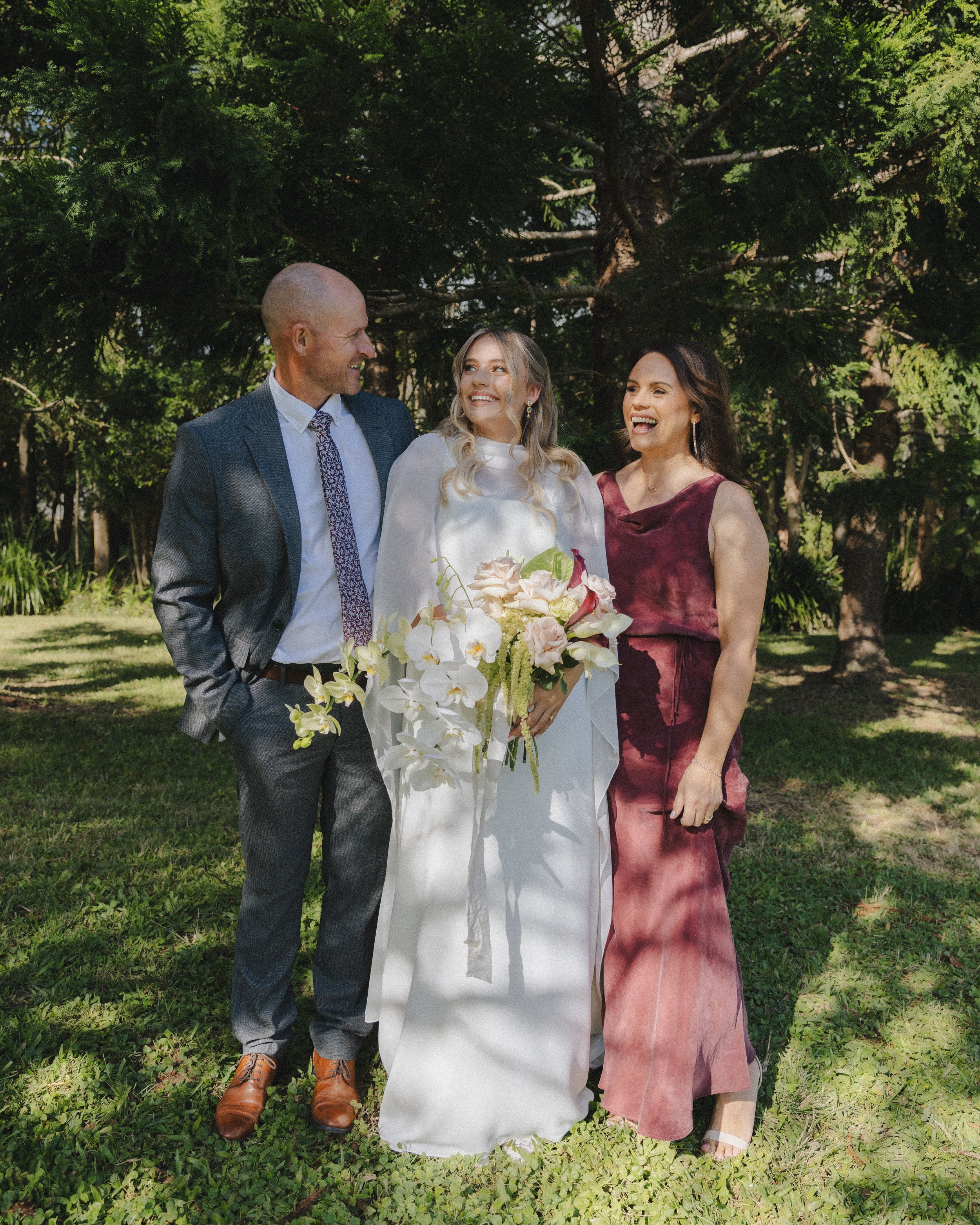 A bride in a white dress holding a bouquet of flowers, standing between a man in a suit and a woman in a burgundy dress, outdoors with green trees in the background, all smiling and looking at each other.