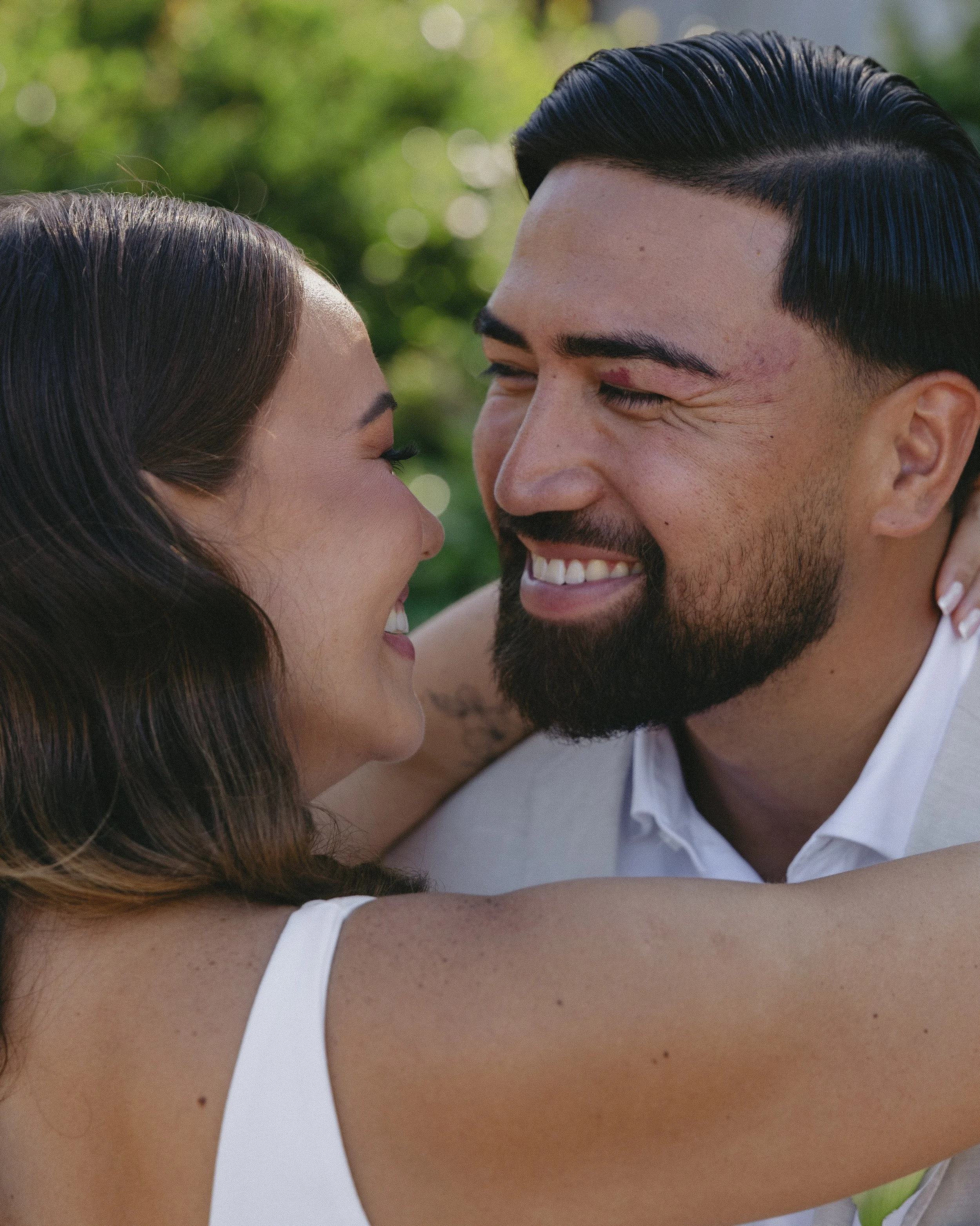 A couple smiling and embracing outdoors with a blurred green background.