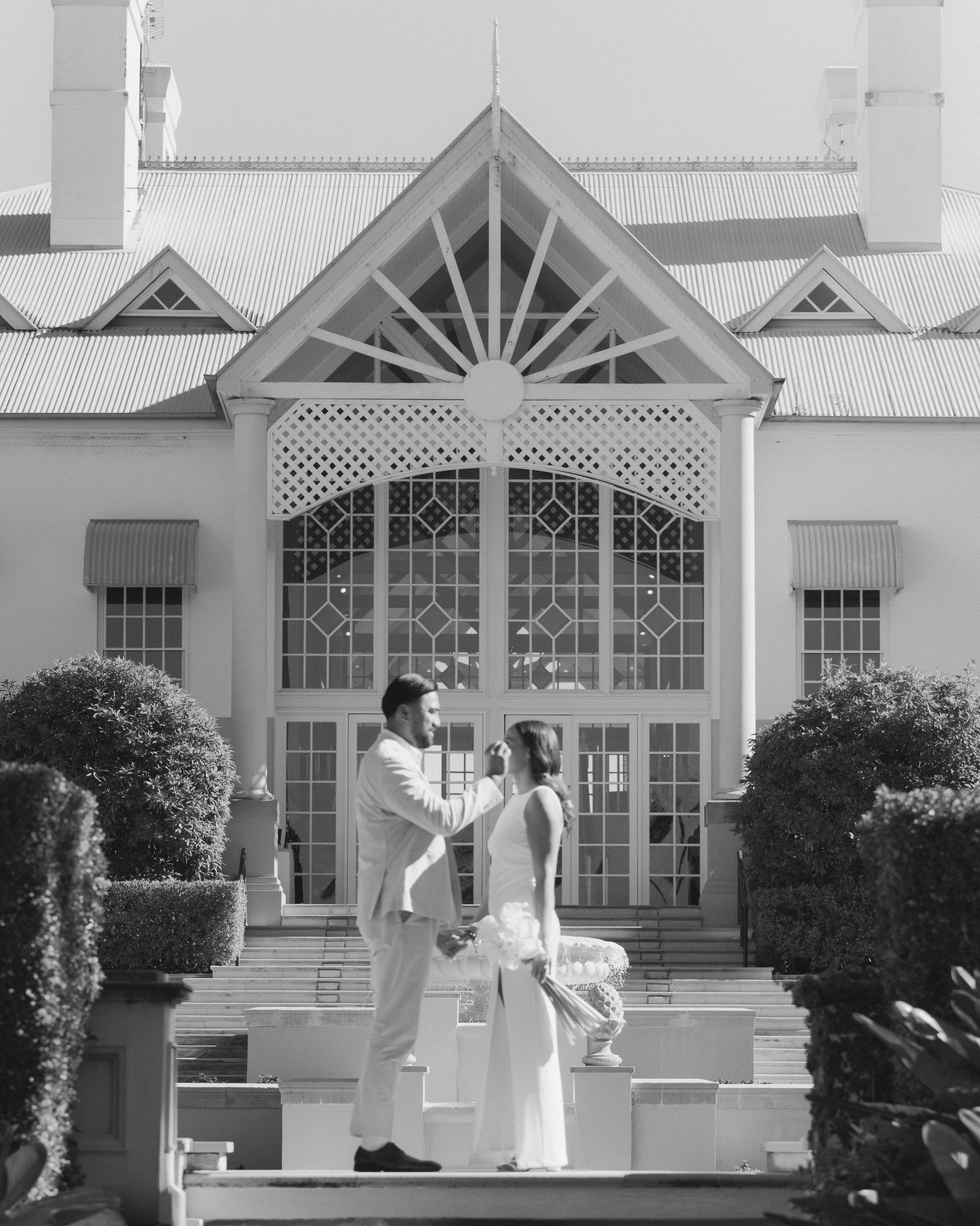 A couple dressed in wedding attire standing on stairs outside a large, ornate building with a high, gabled roof and large windows. The man is feeding the woman a piece of cake, and she is holding a bouquet of flowers. The scene is in black and white.