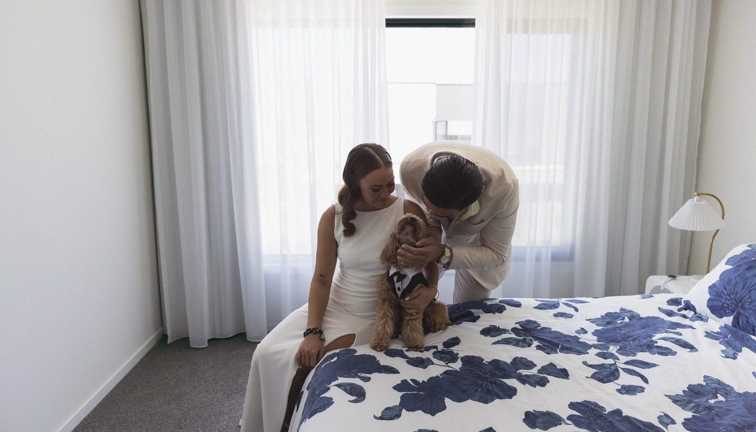 A couple dressed in white interacts with a dog on a bed in a bright, modern bedroom with white curtains and a blue floral bedspread.