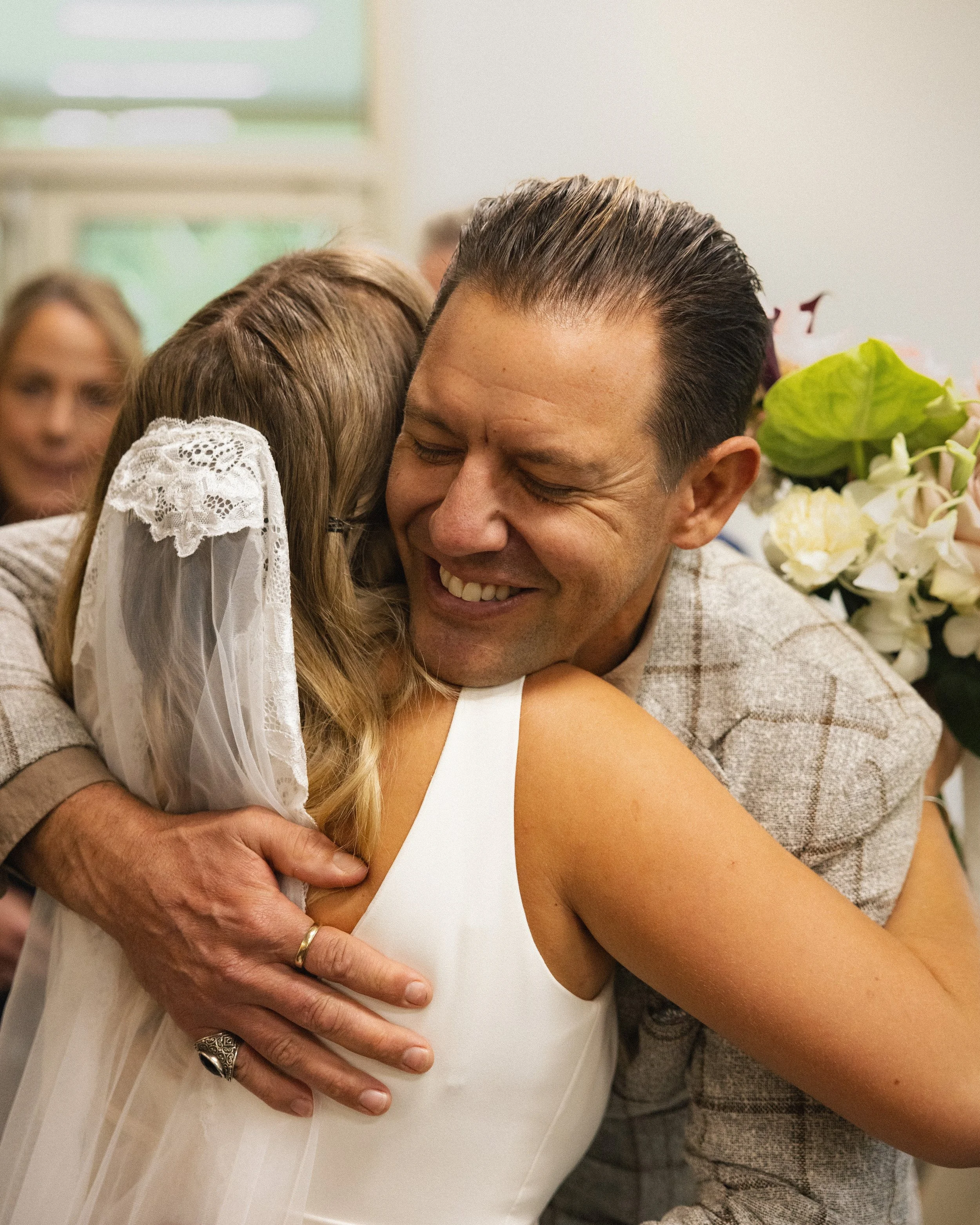 A man and woman sharing a warm hug at a celebration, with the woman wearing a lace-adorned veil and white dress, and the man in a plaid blazer, surrounded by flowers and onlookers.