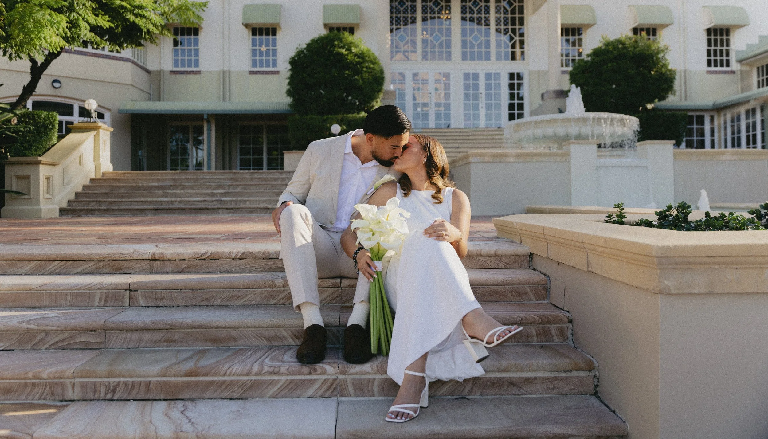 A couple dressed in wedding attire sharing a kiss on the steps of a grand building with a fountain in the background.