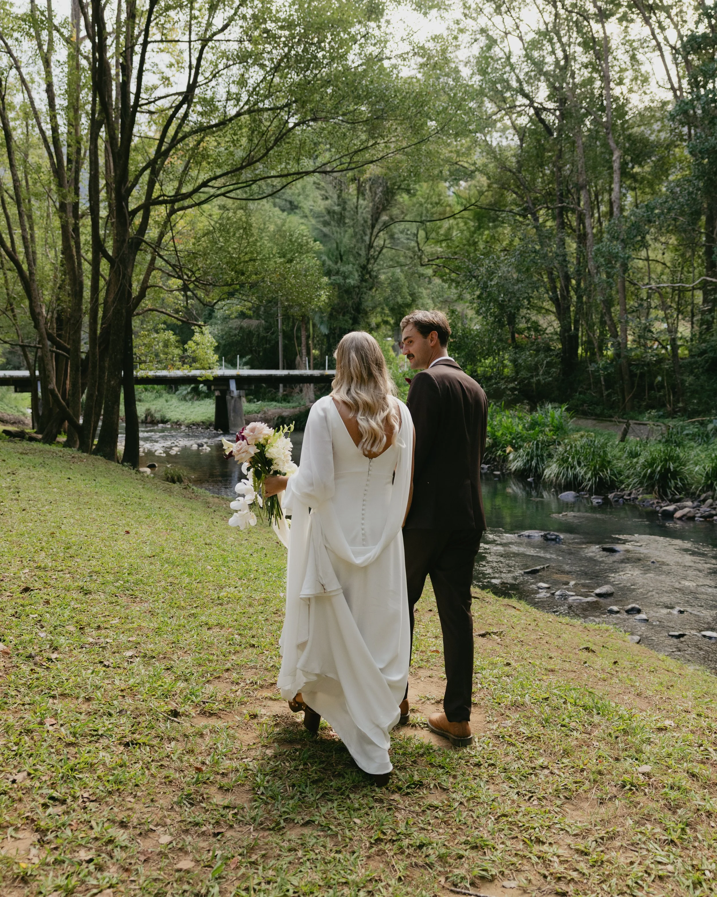 A bride and groom walking together on a grassy path by a creek in a lush, green park with trees and a bridge in the background.