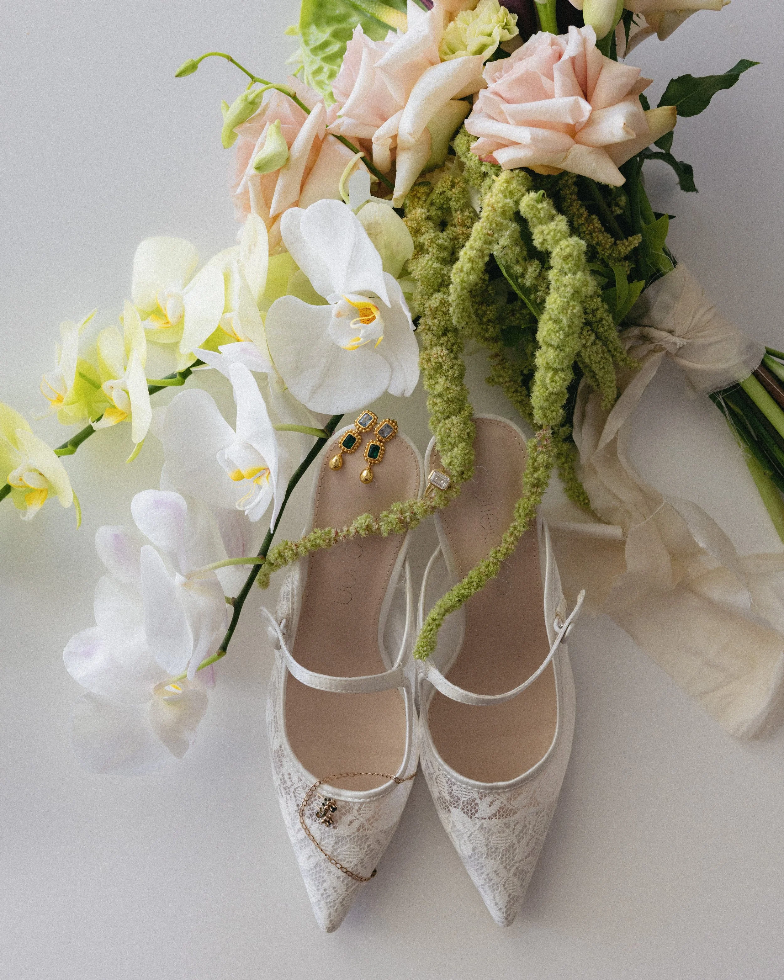 A pair of white lace high heels with jewelry, surrounded by a bouquet of pink roses, white orchids, and greenery.
