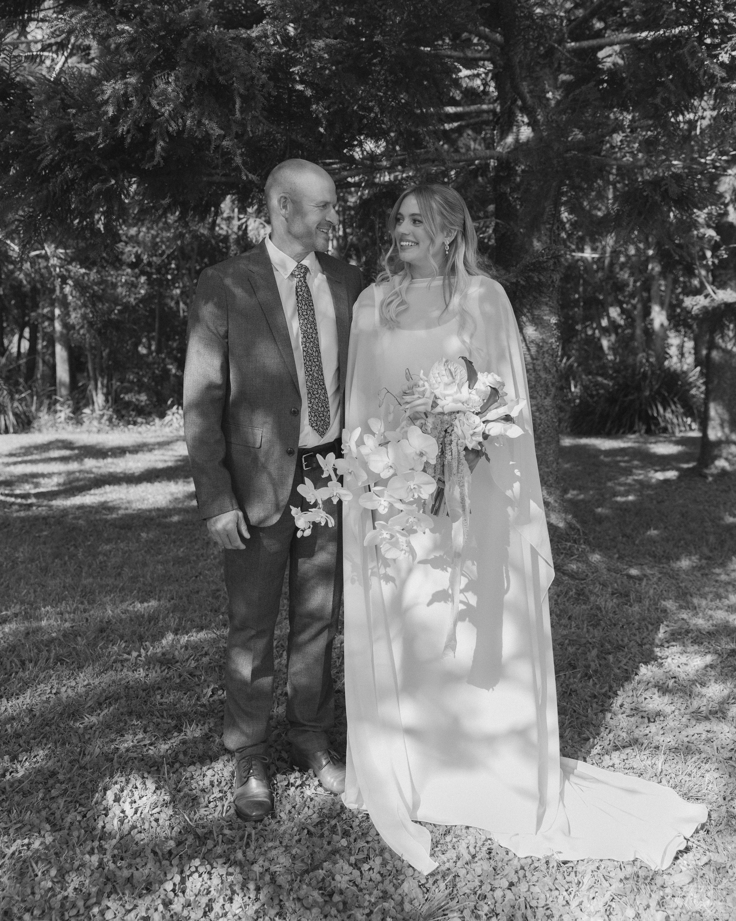 A black-and-white photo of a bride and groom standing outdoors under trees. The bride is holding a bouquet of flowers and wearing a long wedding dress, and the groom is dressed in a suit and tie. They are smiling at each other.