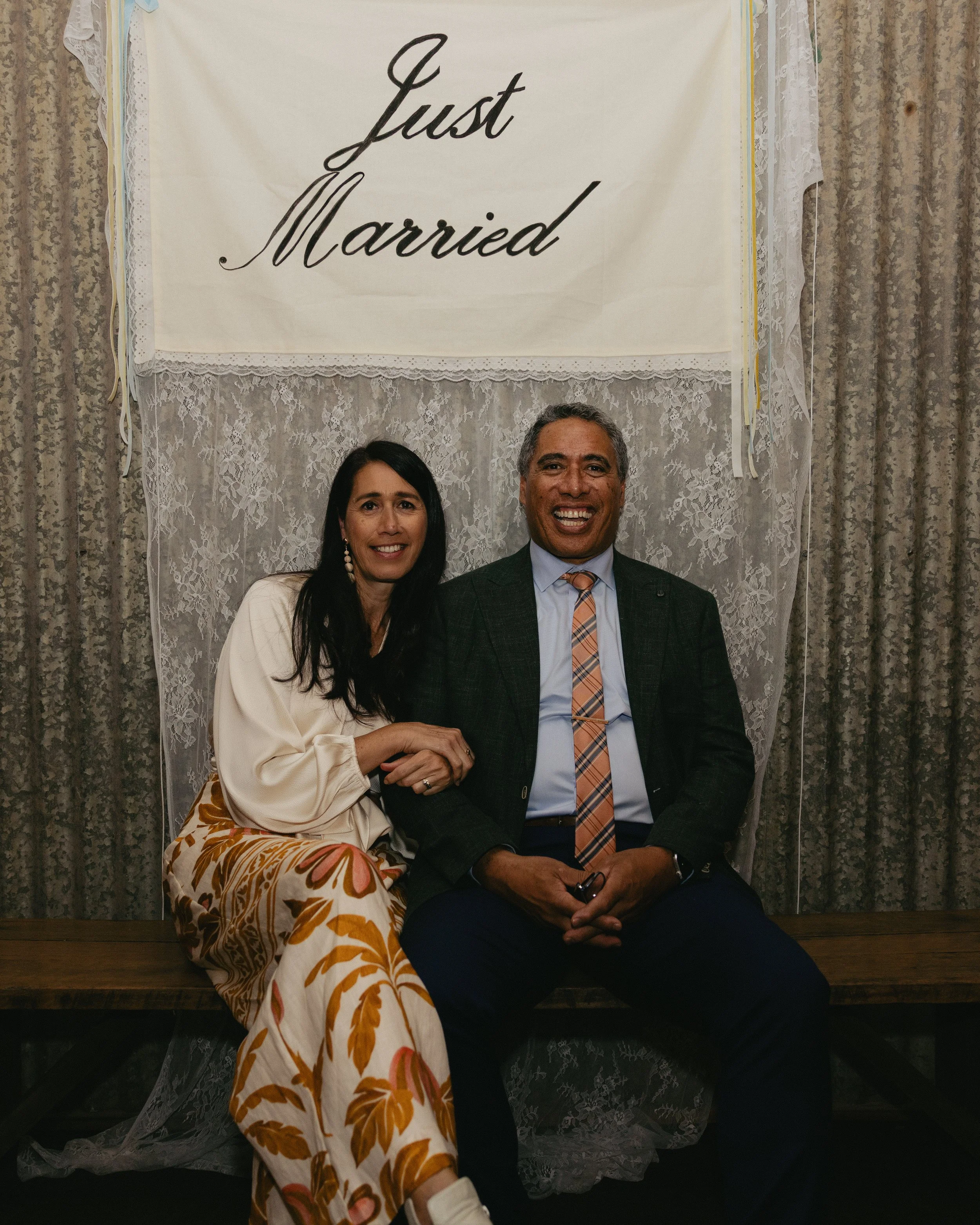 A couple sitting on a wooden bench in front of a lace curtain with a banner that says "Just Married". The woman is wearing a cream blouse and patterned pants, and the man is dressed in a suit with a plaid tie, smiling at the camera.