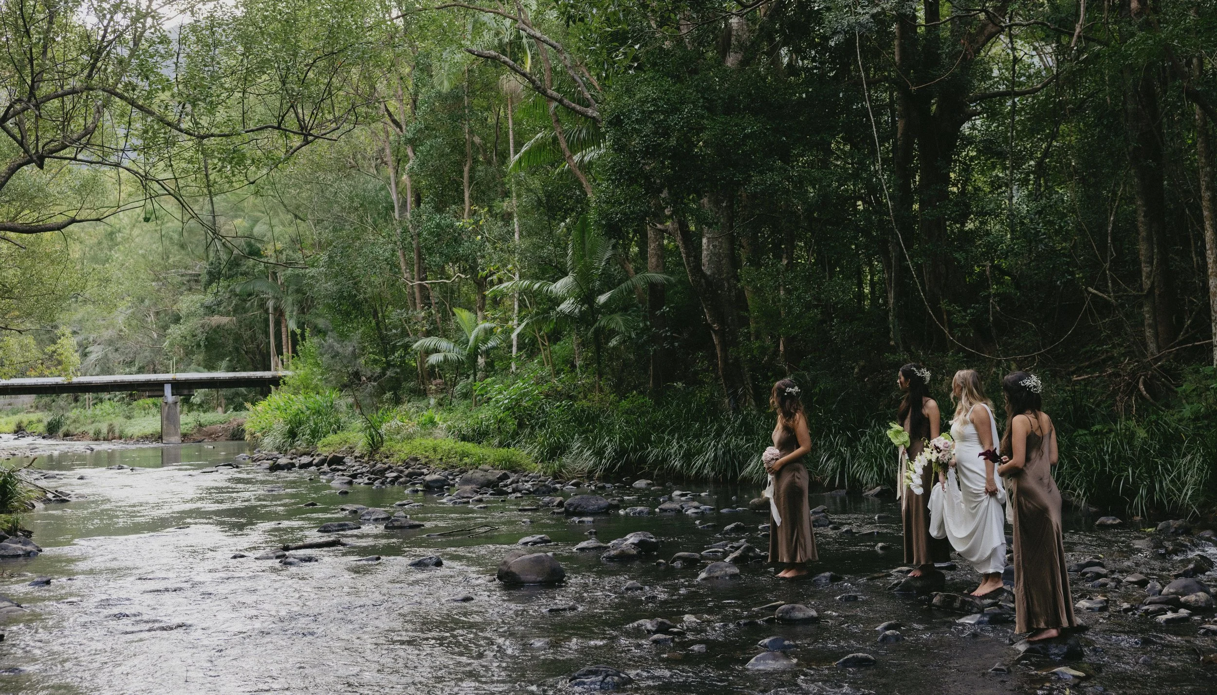 Four women dressed in wedding attire standing in a shallow creek surrounded by lush green trees, with a small bridge visible in the background.