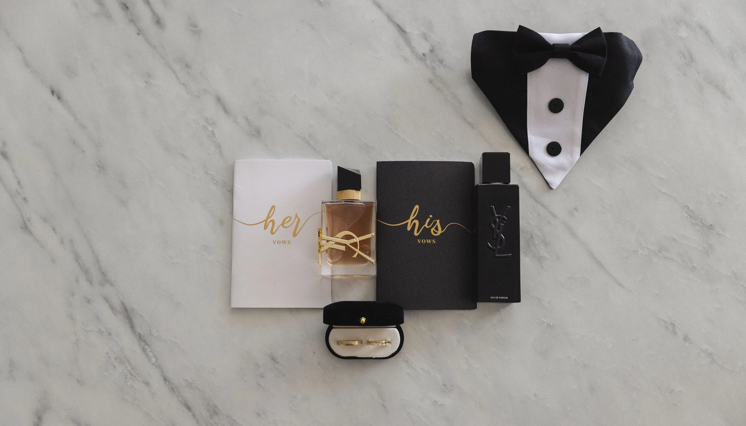 Gift set for wedding vows with two perfume bottles labeled 'her' and 'his', a ring in a black box, gold rings, and black tuxedo-themed attire on a marble surface.