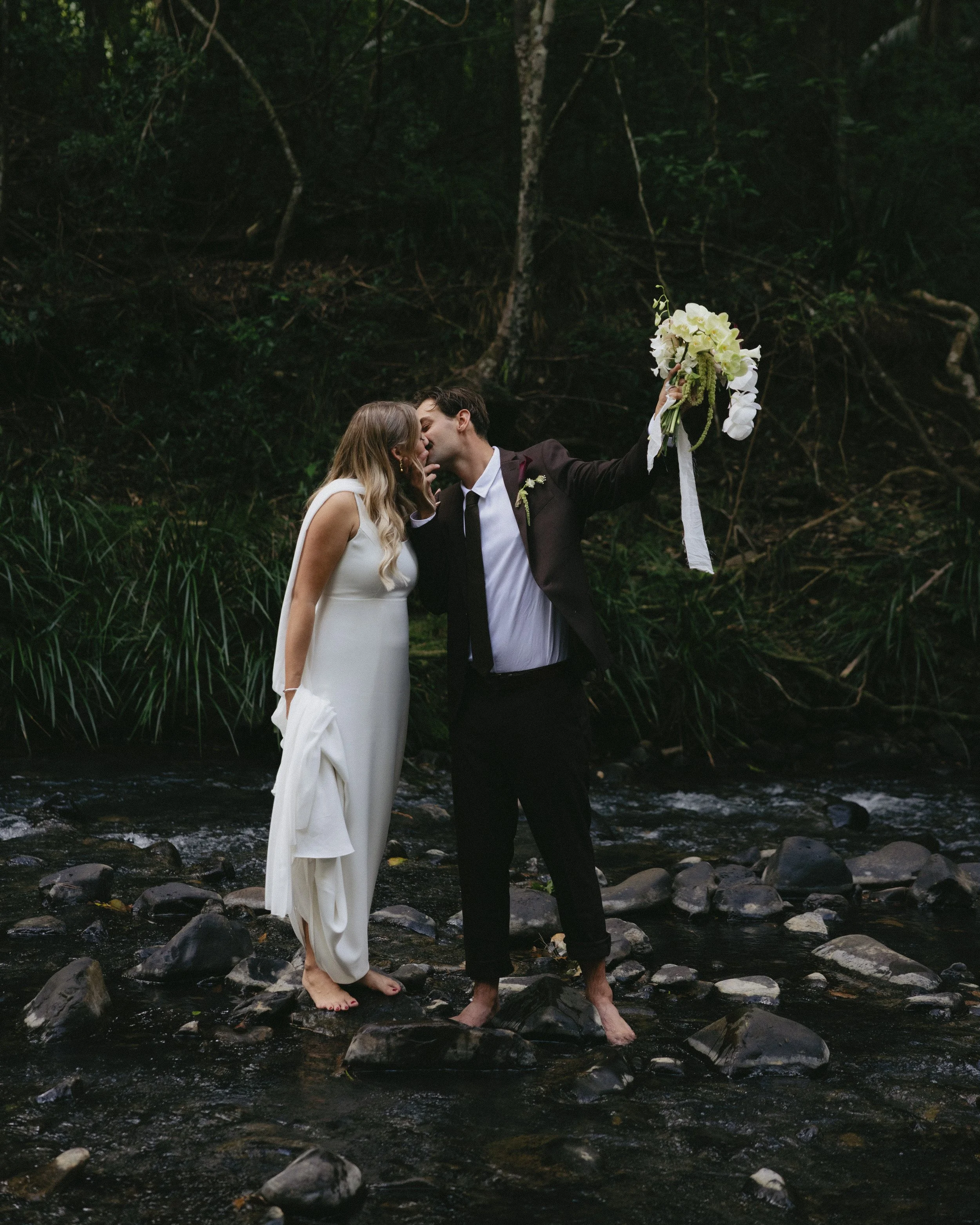 A newlywed couple kissing in a creek surrounded by green foliage, with the groom holding a bouquet of white flowers.